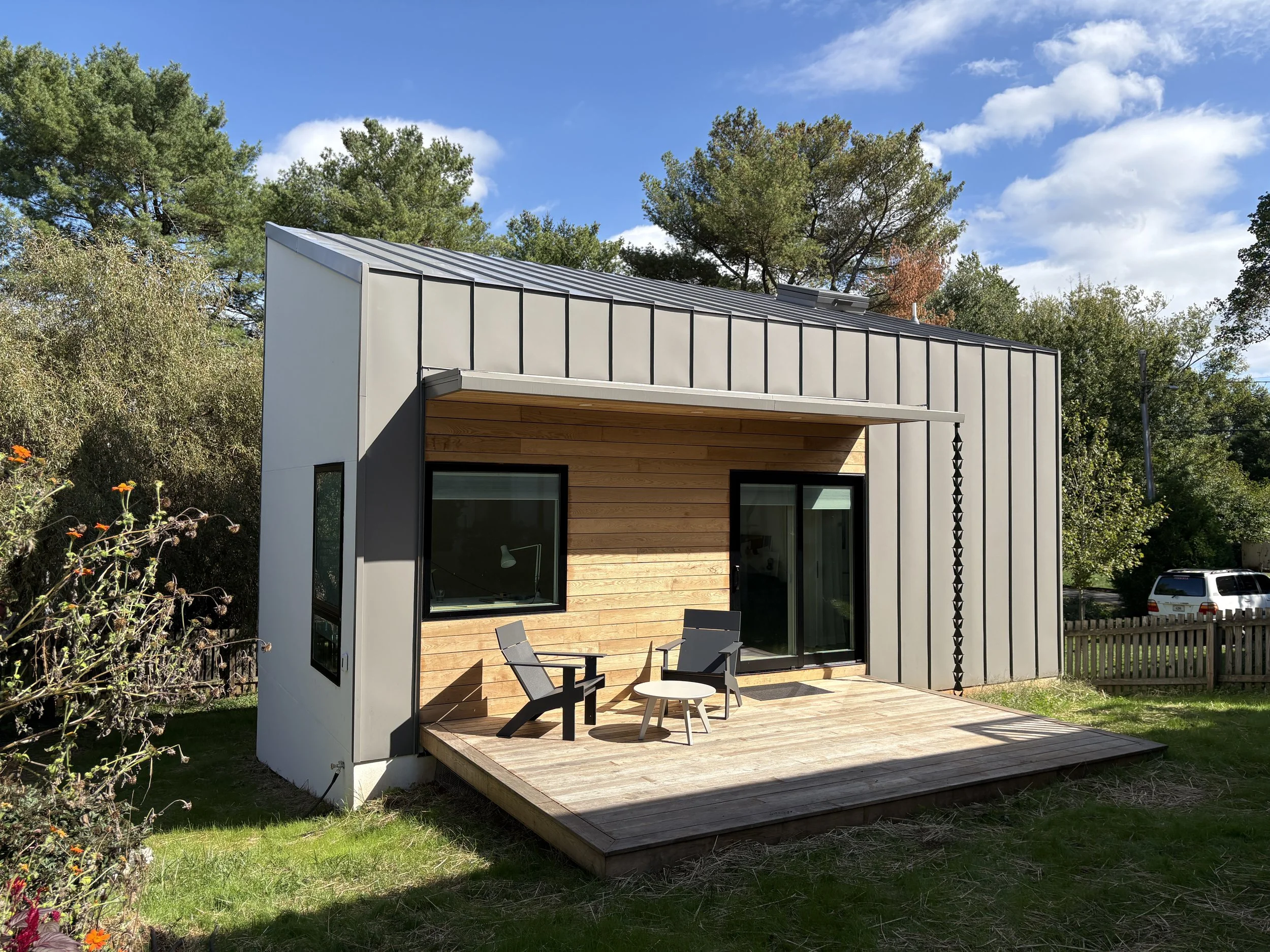 Modern two-story house with metal siding and wooden accents, small raised wooden porch with outdoor chairs, grass yard, surrounded by trees, blue sky with scattered clouds.