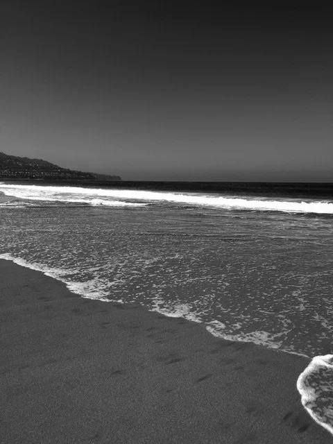 A black and white photo of a calm beach with waves gently crashing onto the shore, and a distant coastline on the horizon.