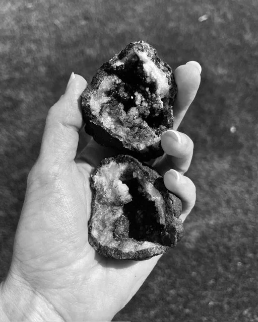 A hand holding two pieces of dark, textured rocks with crystalline formations, against a blurred outdoor background.