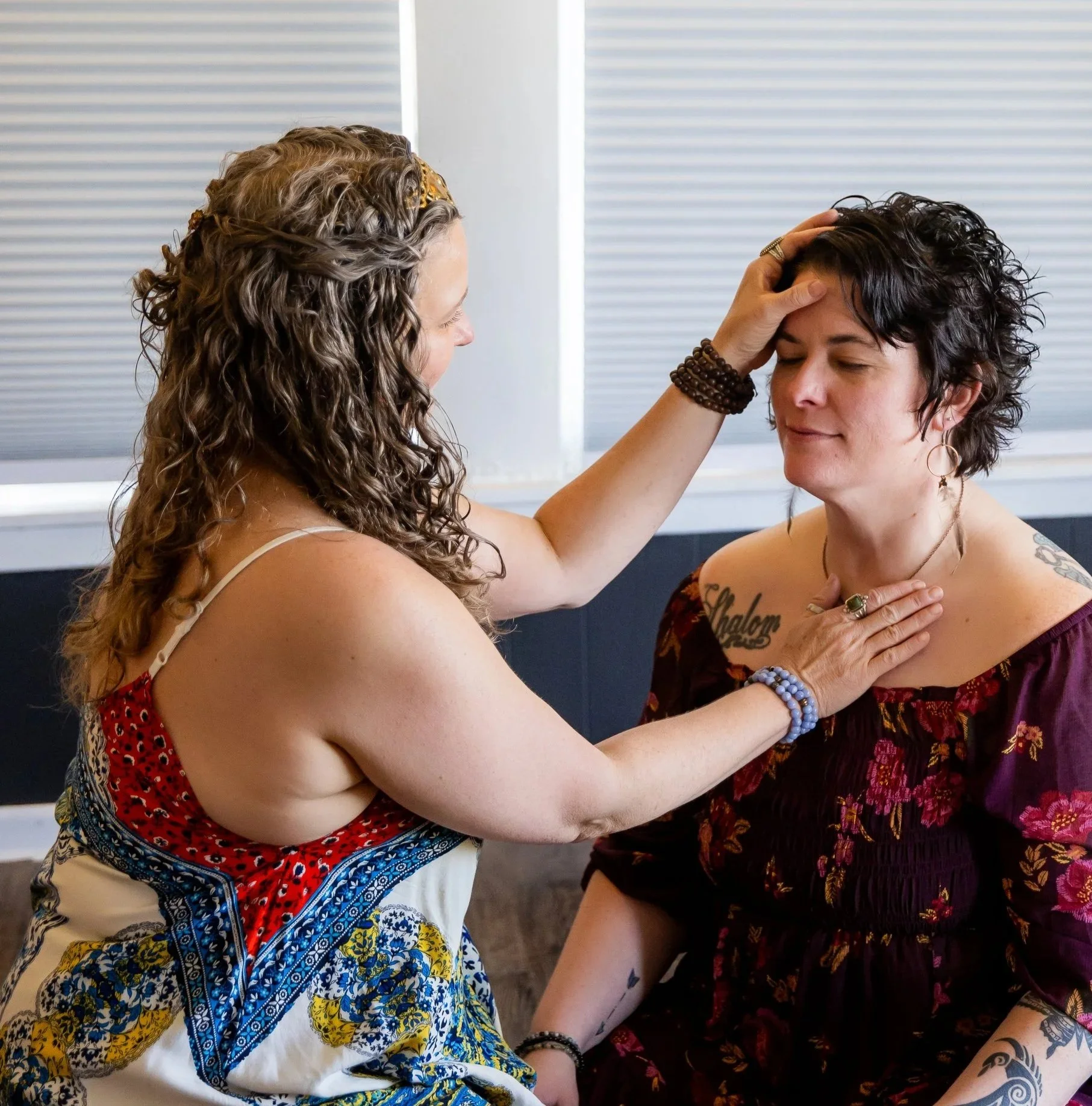 Two women engaged in an emotional moment, one touching the other's forehead and holding her chest, inside a room with closed blinds.