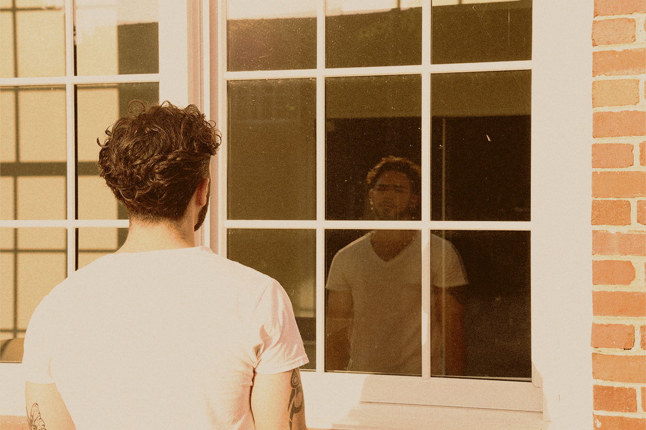 Young man staring at his reflection in a window while deep in thought