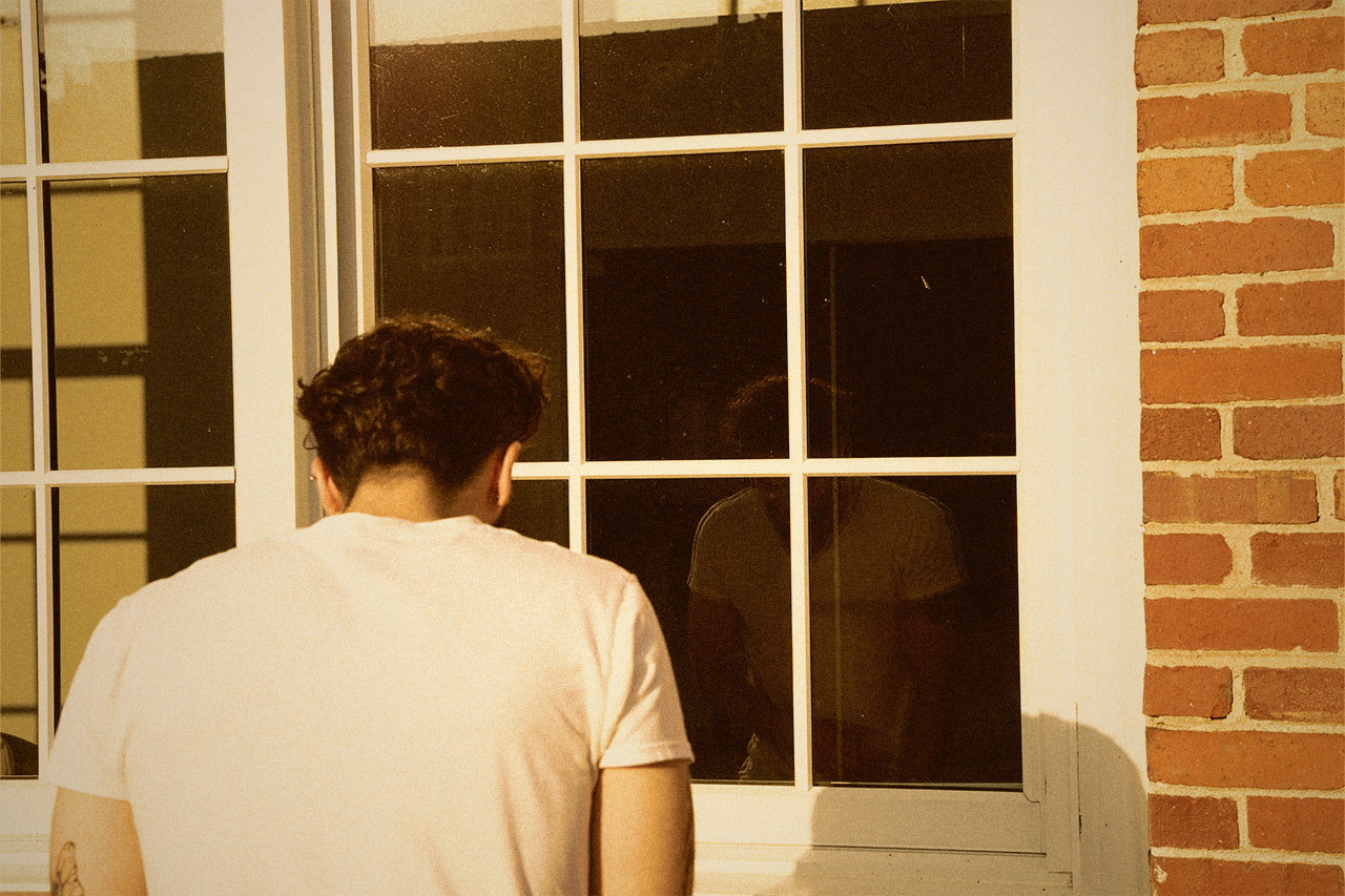 Young man looking down in introspection while standing in front of a window showing his reflection