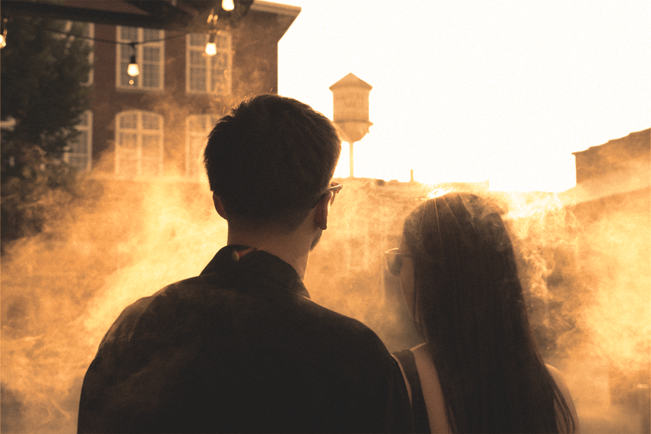 A young man and a young woman with glasses standing together outdoors during sunset, facing away from the camera, with a warm, hazy glow and a brick building in the background.