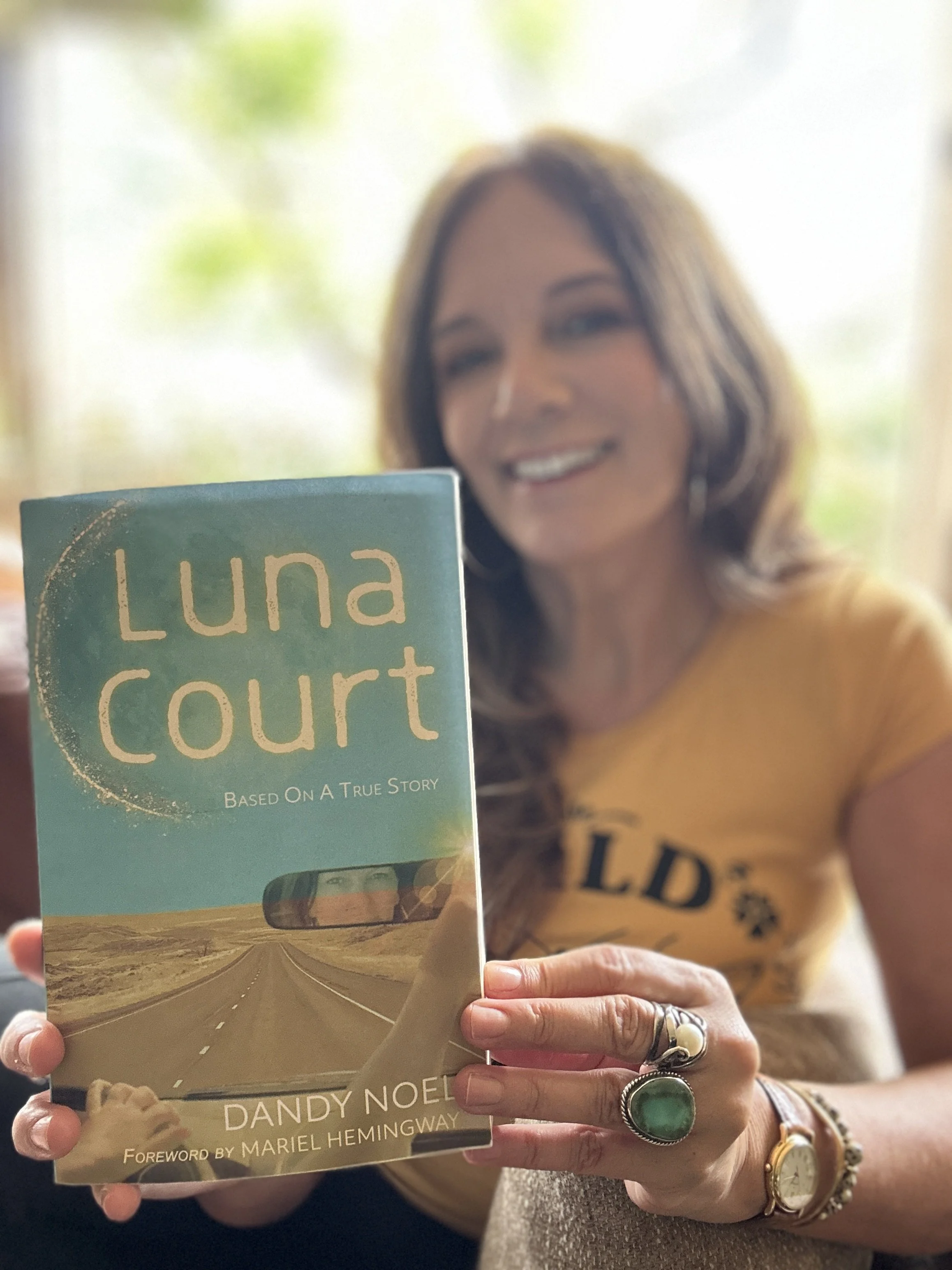 Dandy Noel holding her book titled 'Luna Court' in front of her with a desert road scene on the cover, smiling at the camera.