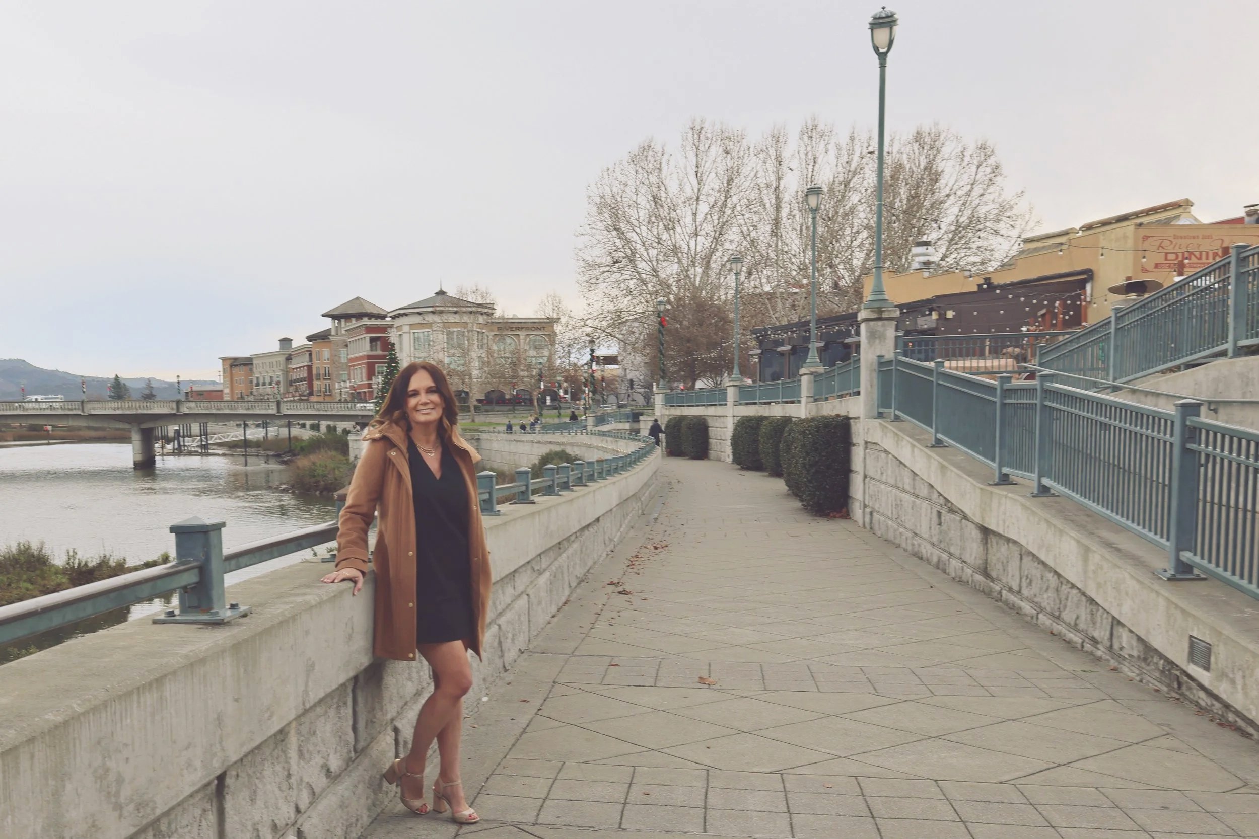 Dandy Noel wearing a black dress and tan coat standing on a riverside walkway with a city skyline and leafless trees in the background.