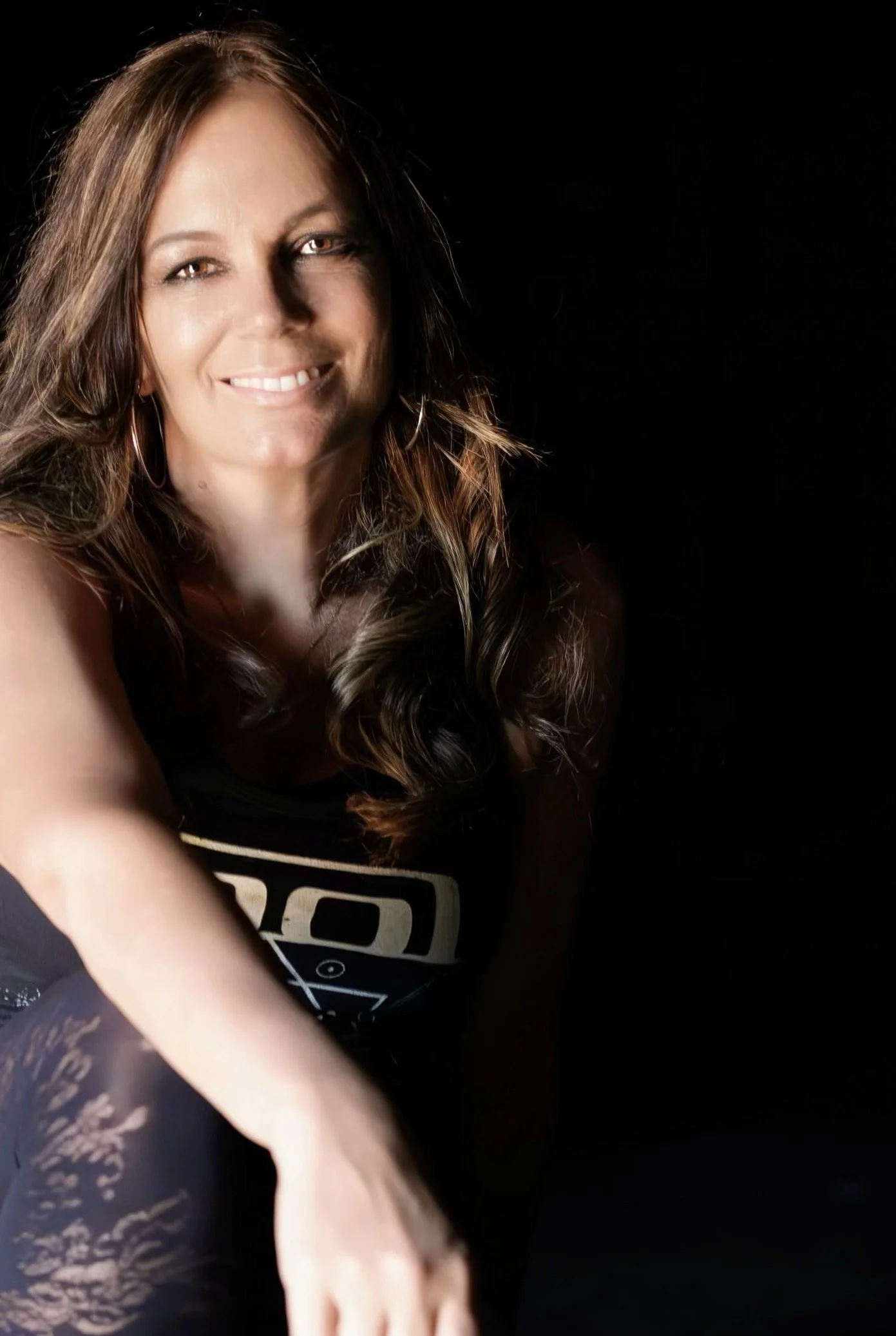 Portrait of Dandy Noel smiling, with long wavy brown hair, against a dark background.