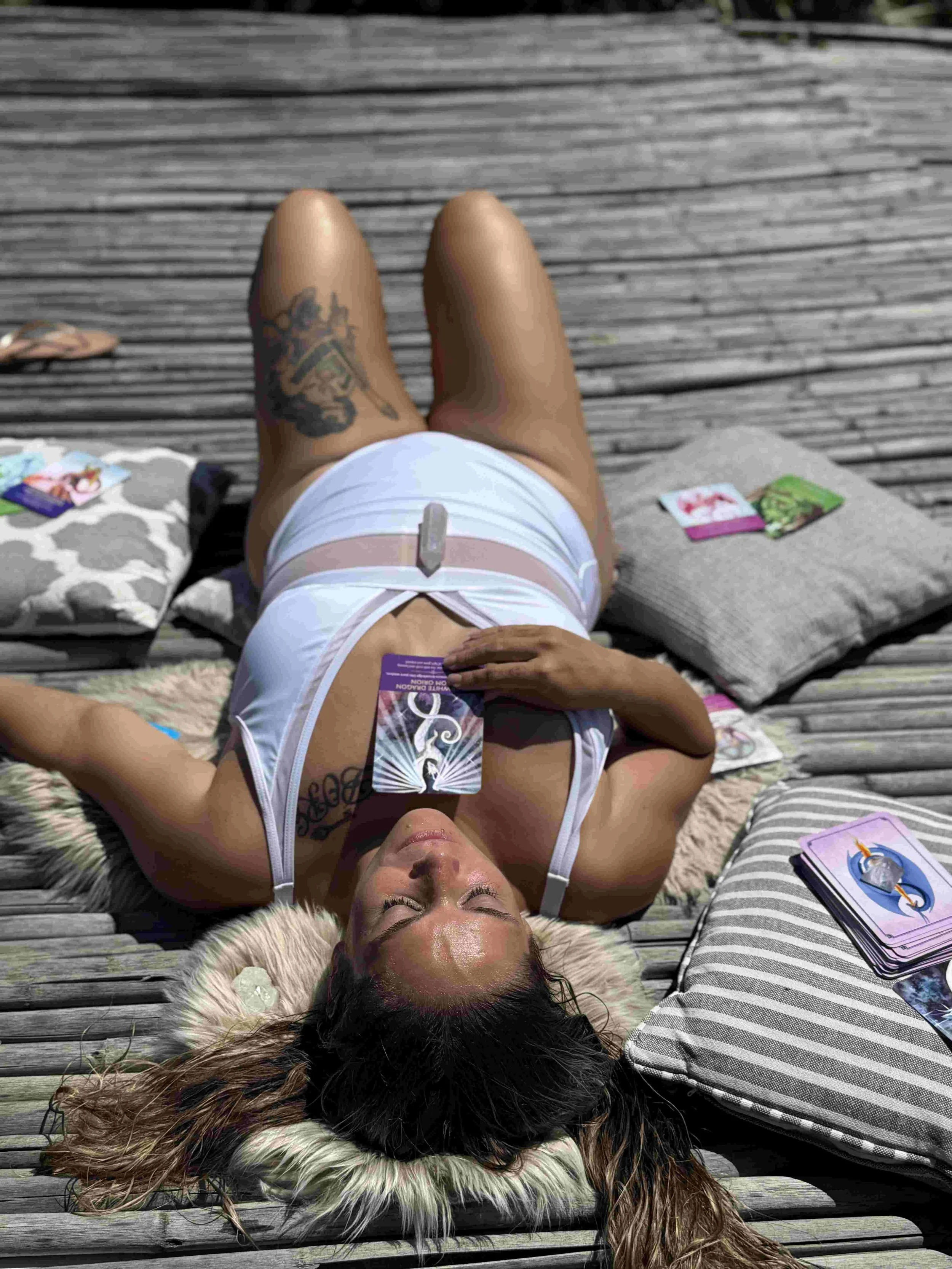 A woman with long hair lying on a fur-lined pillow on a wooden deck, holding tarot cards, with tarot card decks and cushions around her.