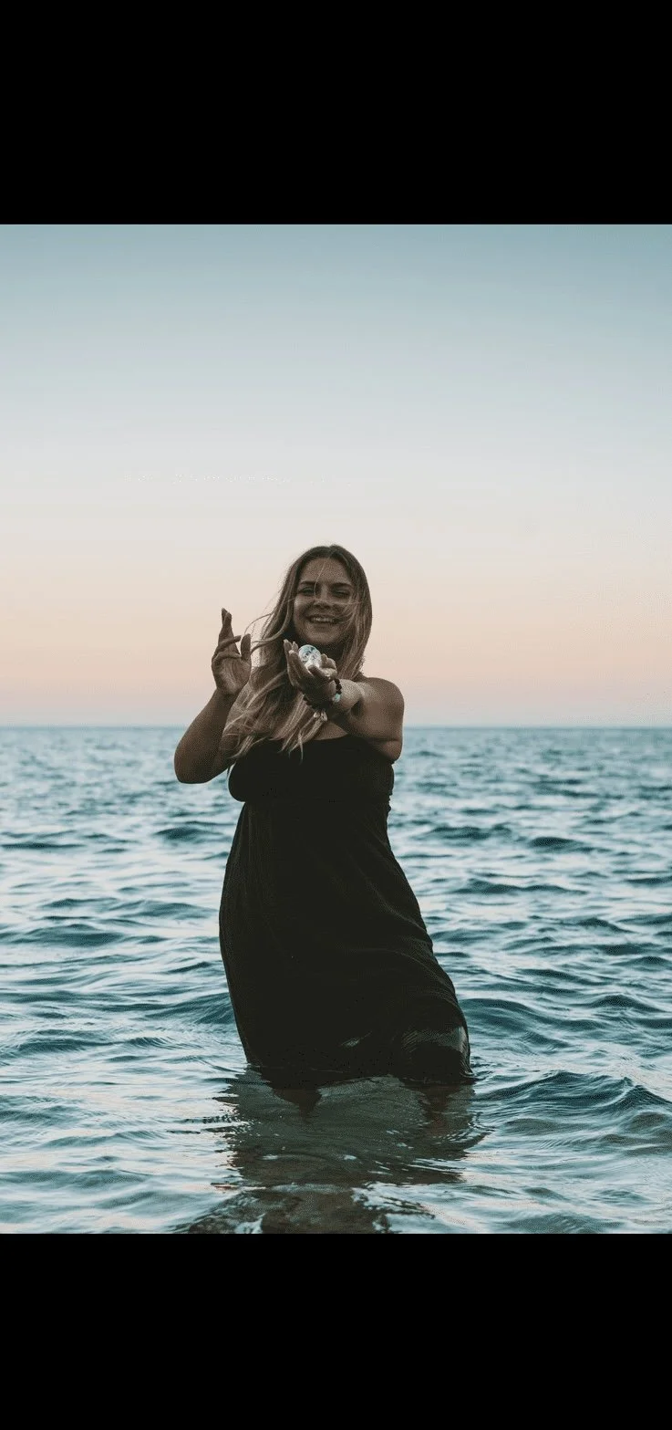 A woman in a black dress standing in the ocean during sunset, smiling and holding water.