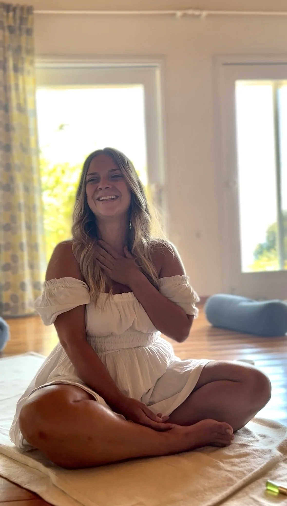 A woman sitting cross-legged on a yoga mat, smiling and touching her neck, in a room with large windows and natural light.