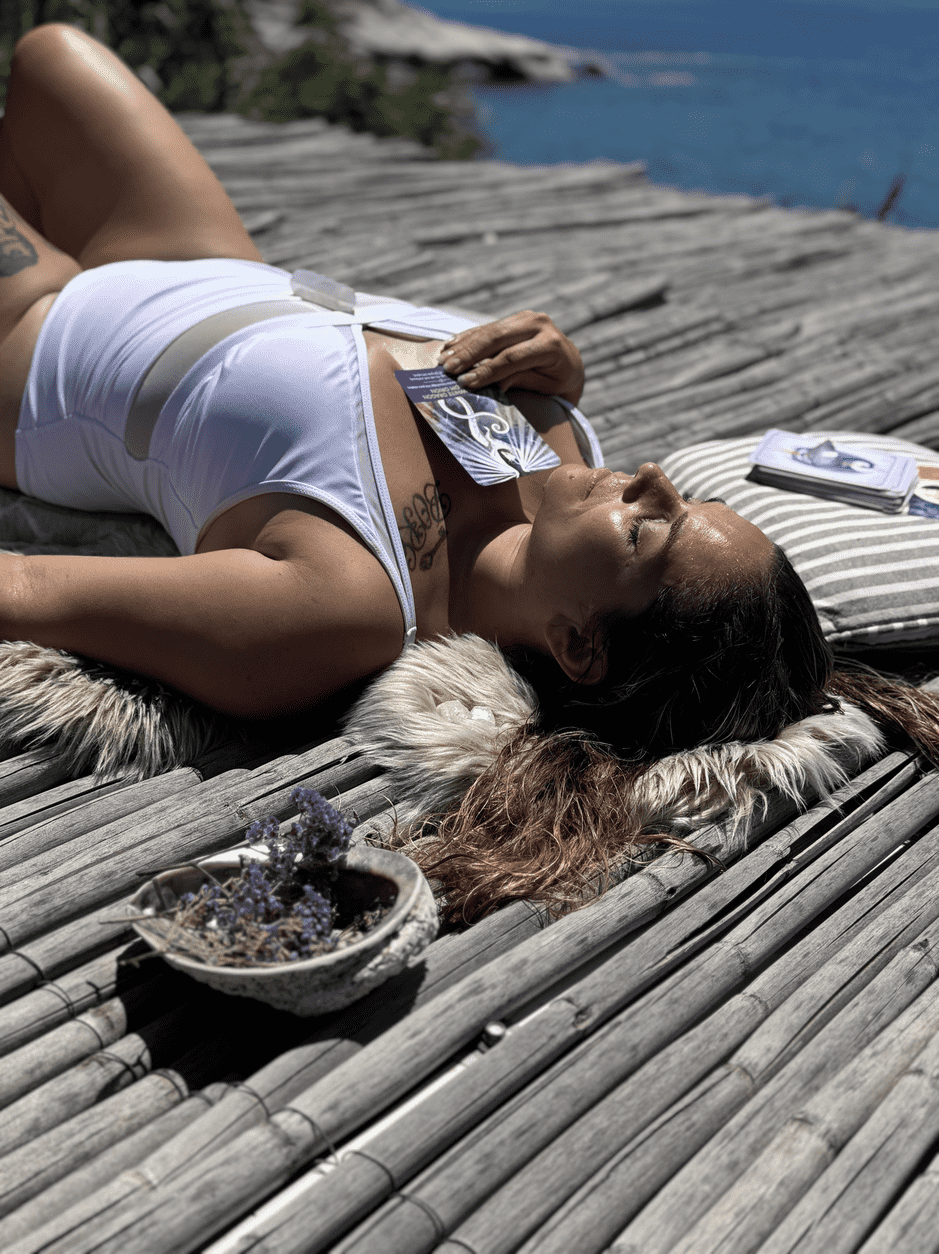 Woman lying on a bamboo dock near water, sunbathing with a tarot card  on her white swimsuit, surrounded by a bowl of purple flowers and a striped pillow.