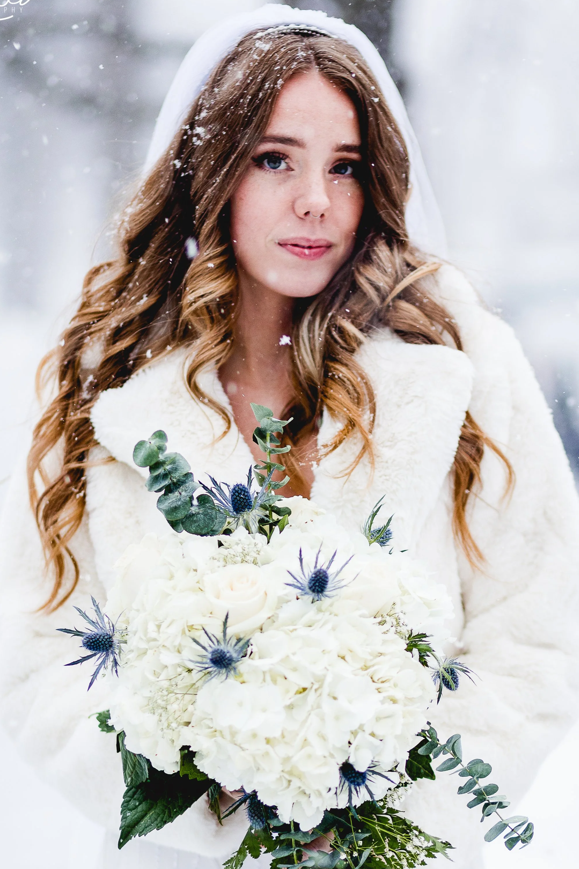 A woman with long, wavy red hair, dressed in a white coat, holding a bouquet of white and blue flowers, standing outdoors in a snowy setting.