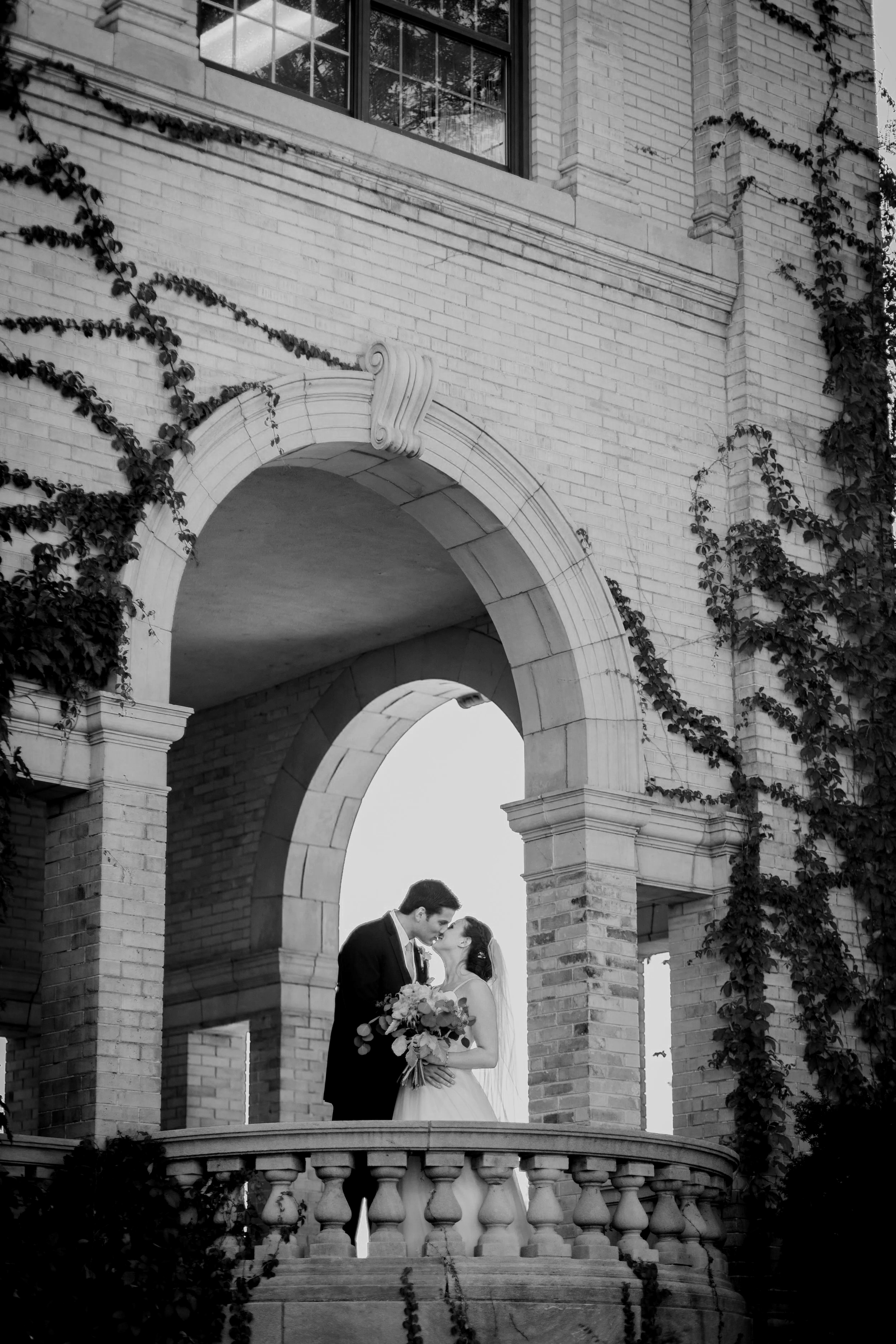 A bride and groom sharing a kiss on a stone balcony with an arched entryway, surrounded by ivy-covered brick walls, during a wedding.