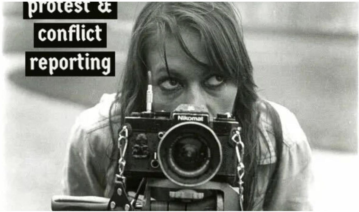 Black and white photo of a woman with long hair taking a self-portrait with a Nikon camera, with a sign in the background reading 'protest & conflict reporting'.