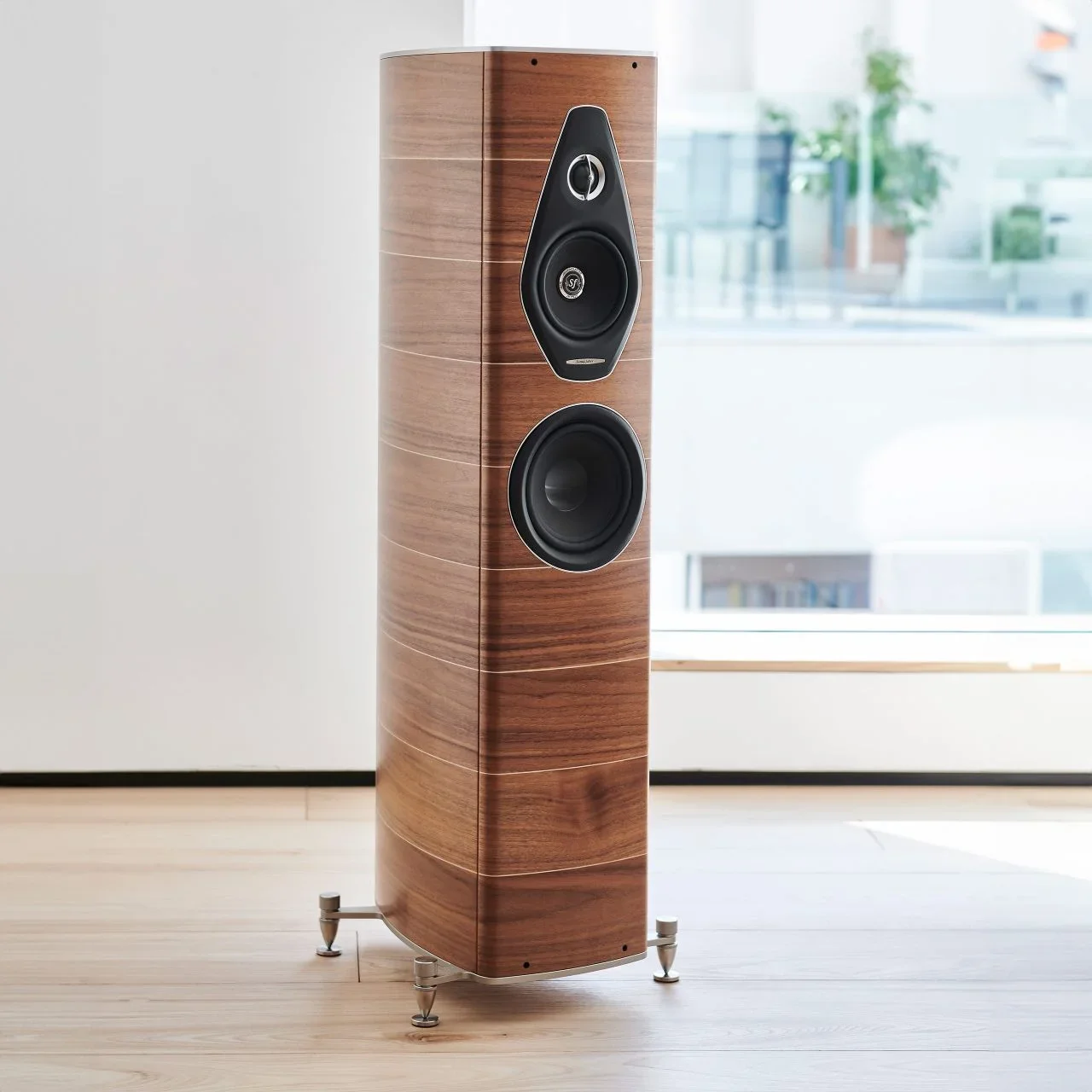 A tall floor-standing speaker with a wood grain finish, positioned indoors on a light wooden floor near large windows with views of trees and buildings outside.