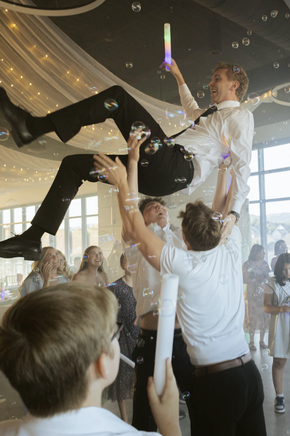 Groomsmen lifting the groom in celebration as bubbles float across a sunlit reception
