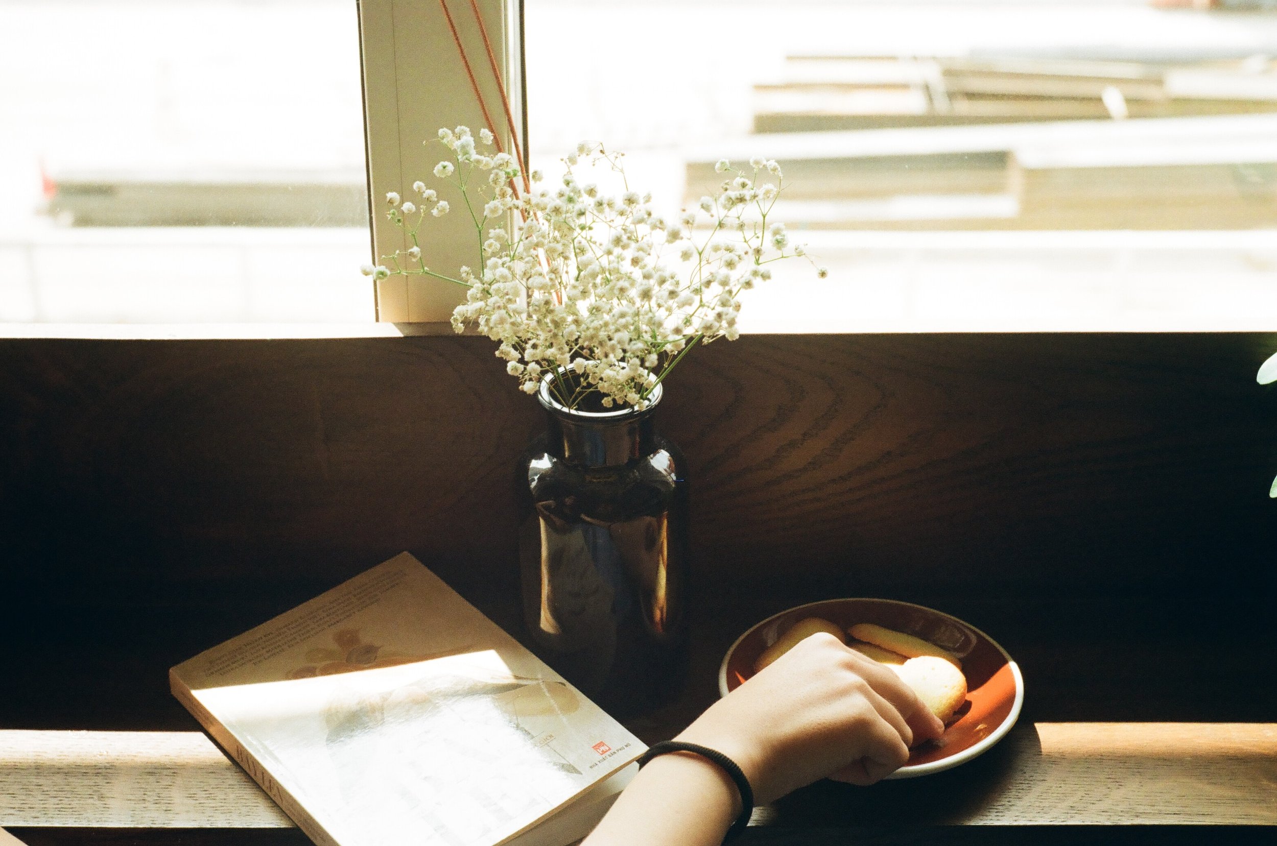 A person sitting at a wooden table with a gentle background, holding a piece of food on a small orange and white plate. Nearby on the table, there is a book and a dark glass vase with white baby's breath flowers. Sunlight filters through a nearby window illuminating the scene.