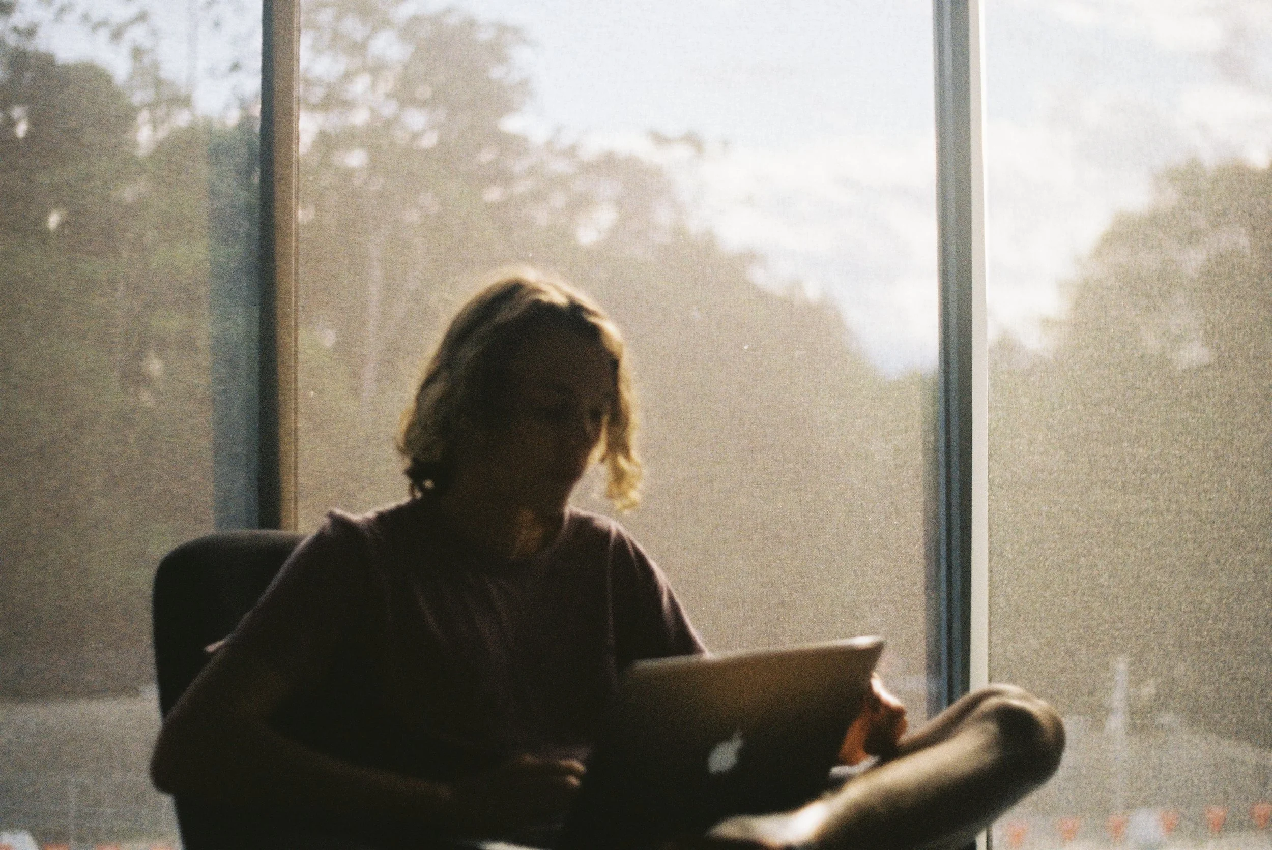 Silhouette of a person with blonde hair sitting at a table near a large window, with sunlight streaming in and trees visible outside.