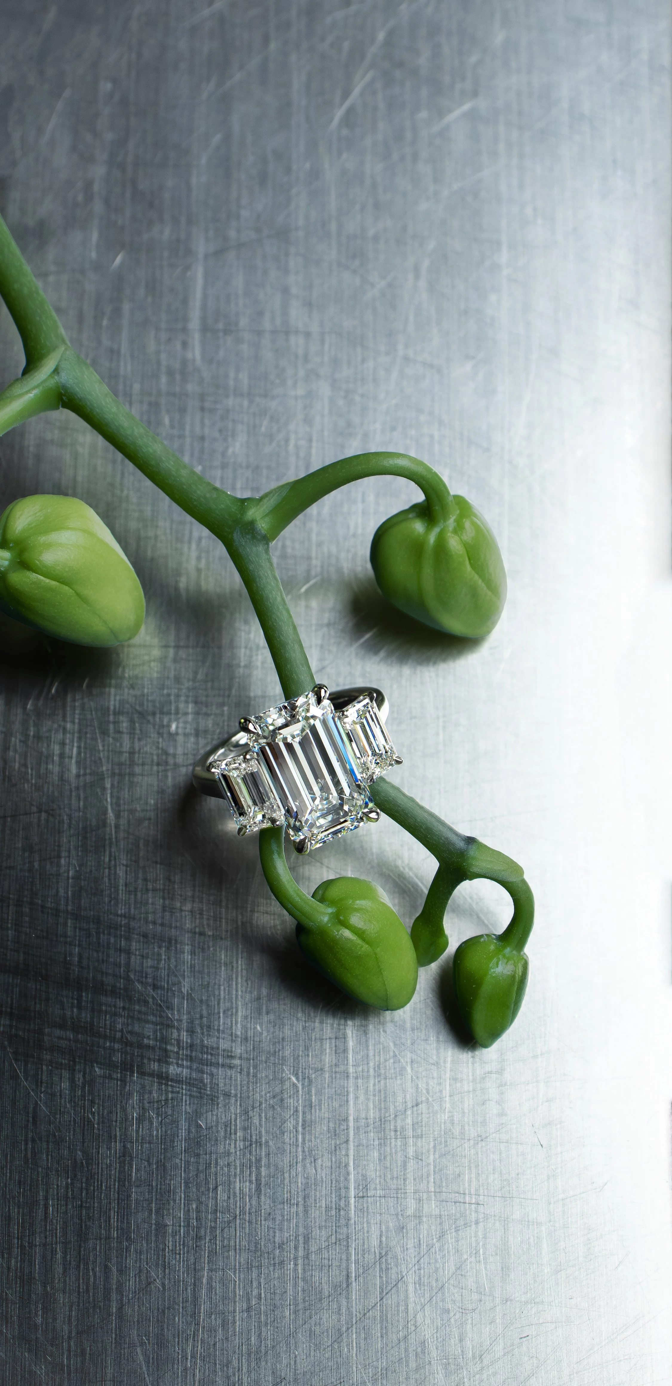 Platinum three-stone ring with an elongated emerald-cut center diamond and matching side stones, photographed with green orchid stems.