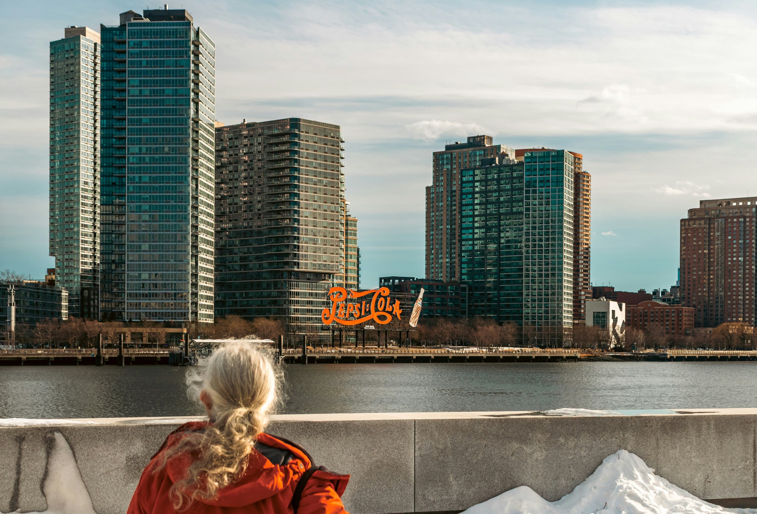 An elderly person with gray hair sitting by a river, looking at tall modern buildings in the distance. The buildings include glass skyscrapers and a sign with Pepsi-Cola on the riverfront. There is snow on the ground and a cloudy sky above.