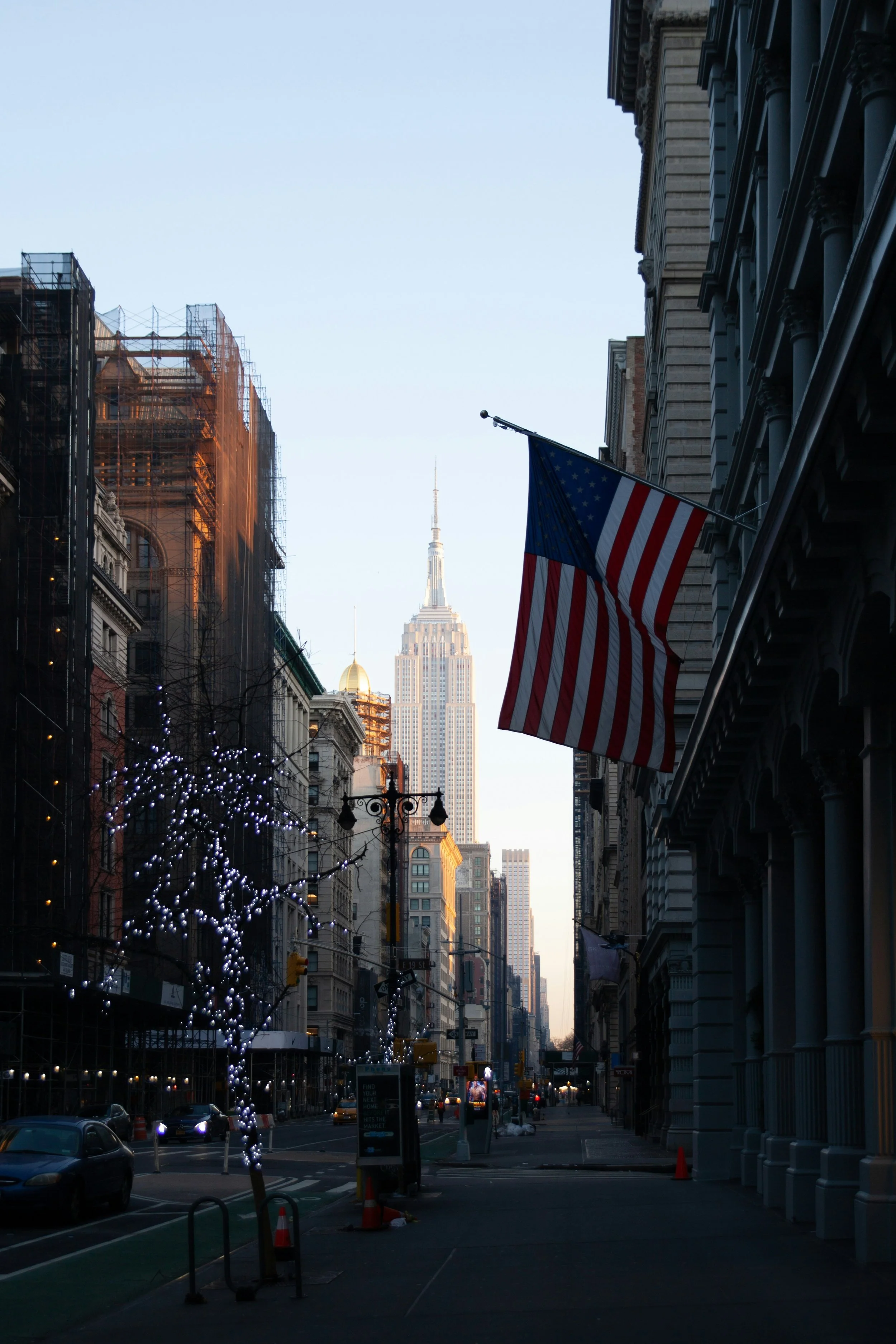 City street scene with tall buildings, American flags, and Empire State Building in the background under a clear sky, with some Christmas lights on trees and street lamps.