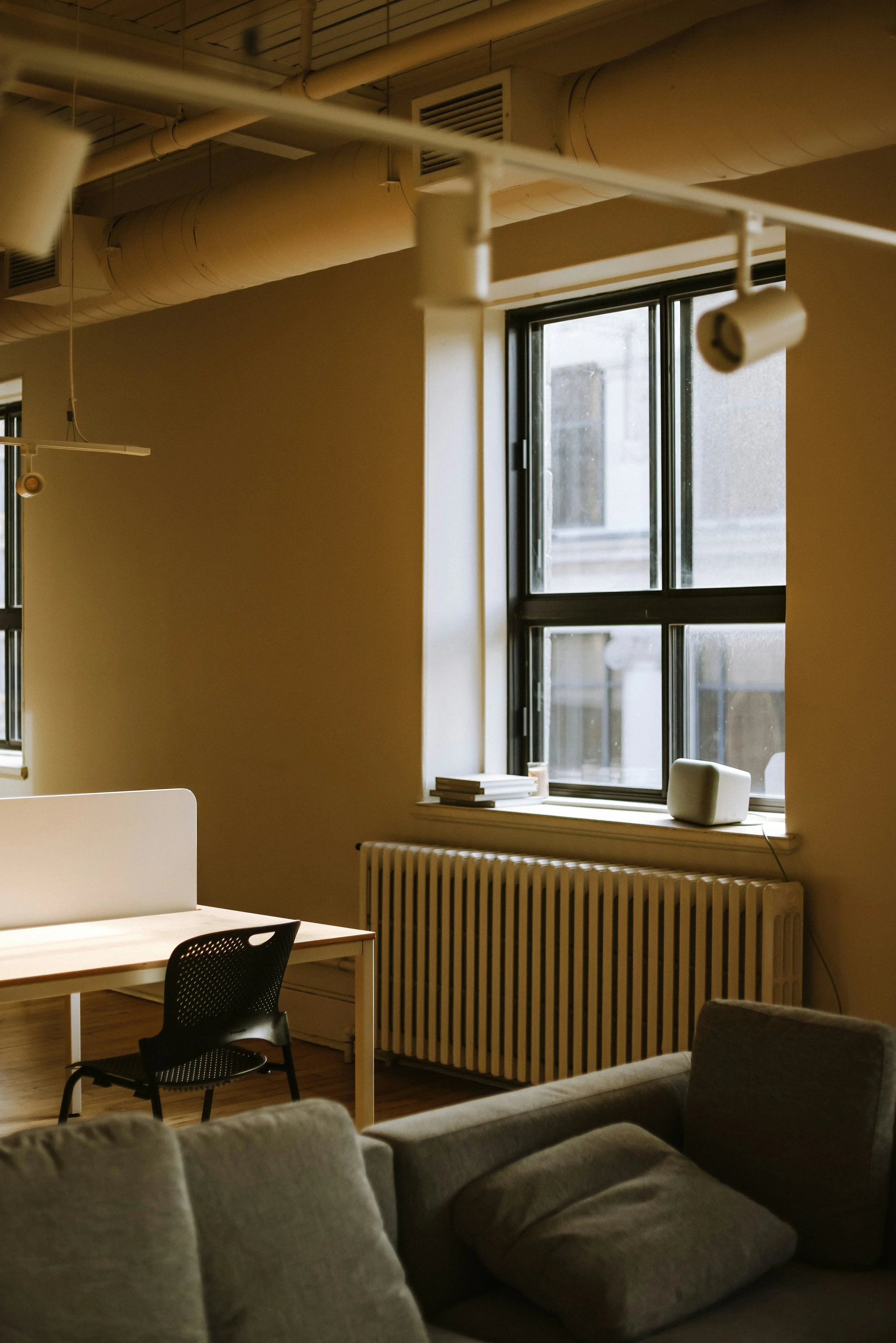 Interior view of an office or living space with large windows, a radiator, a table, a chair, and a sofa, illuminated by natural light.