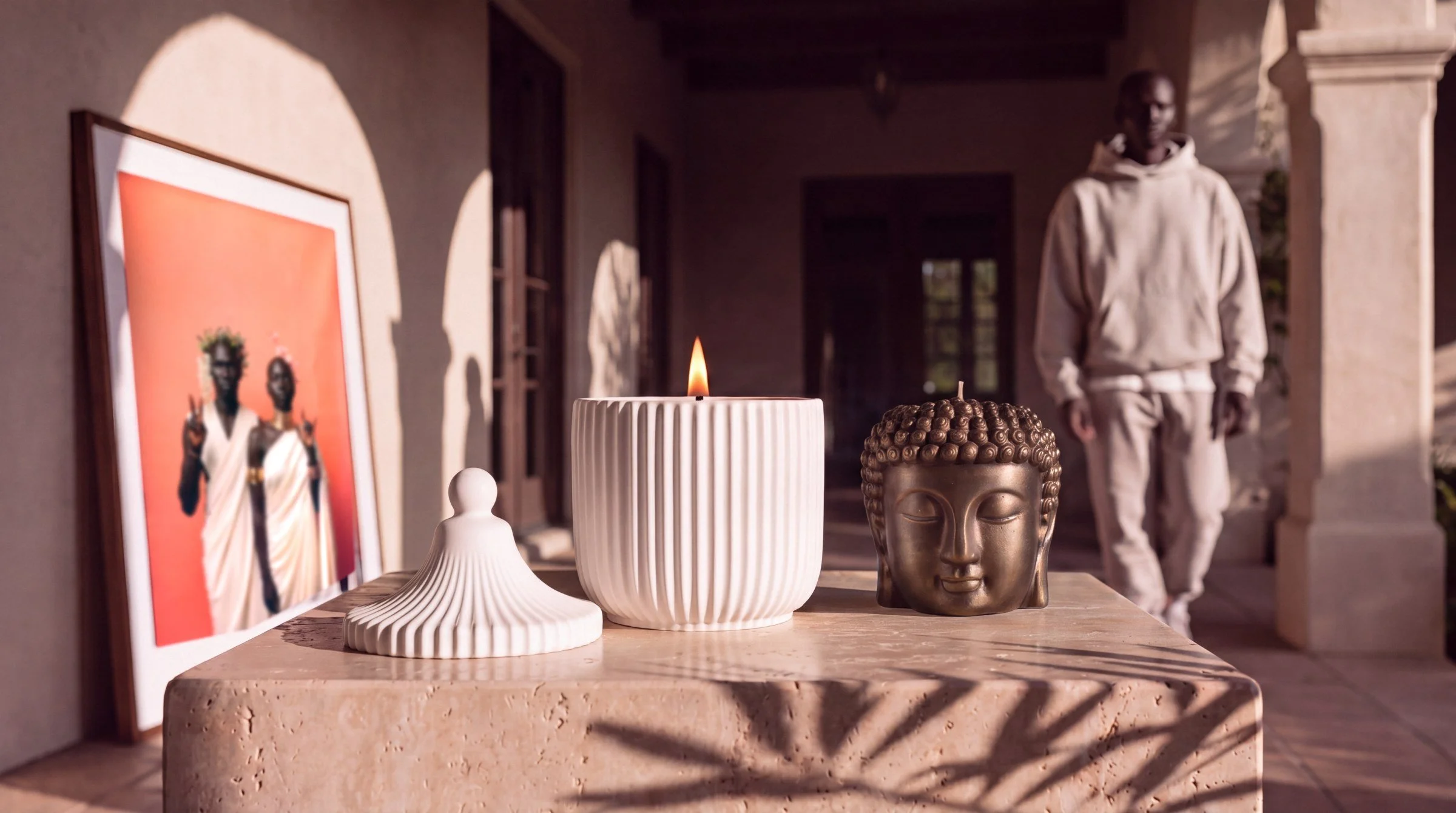 A lit Belaire candle and a bronze Buddha candle on travertine, with the Peace and Love print leaning on the wall behind