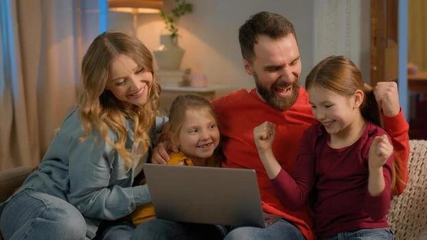 A happy family of four watching a laptop together on a couch, with the father and daughter showing excitement.
