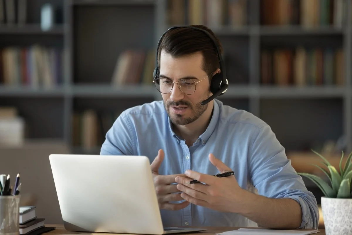 A man wearing a headset with a microphone sitting at a desk in front of a laptop performing laptop remote support.