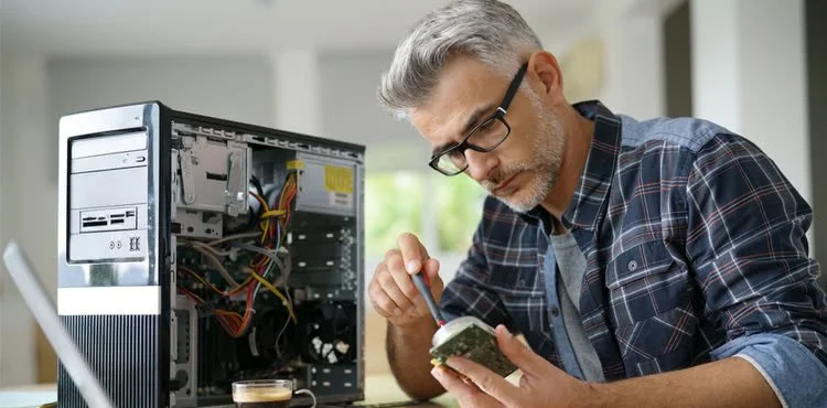 Man repairing a computer tower, holding a circuit board and using a tool, next to an opened desktop case.