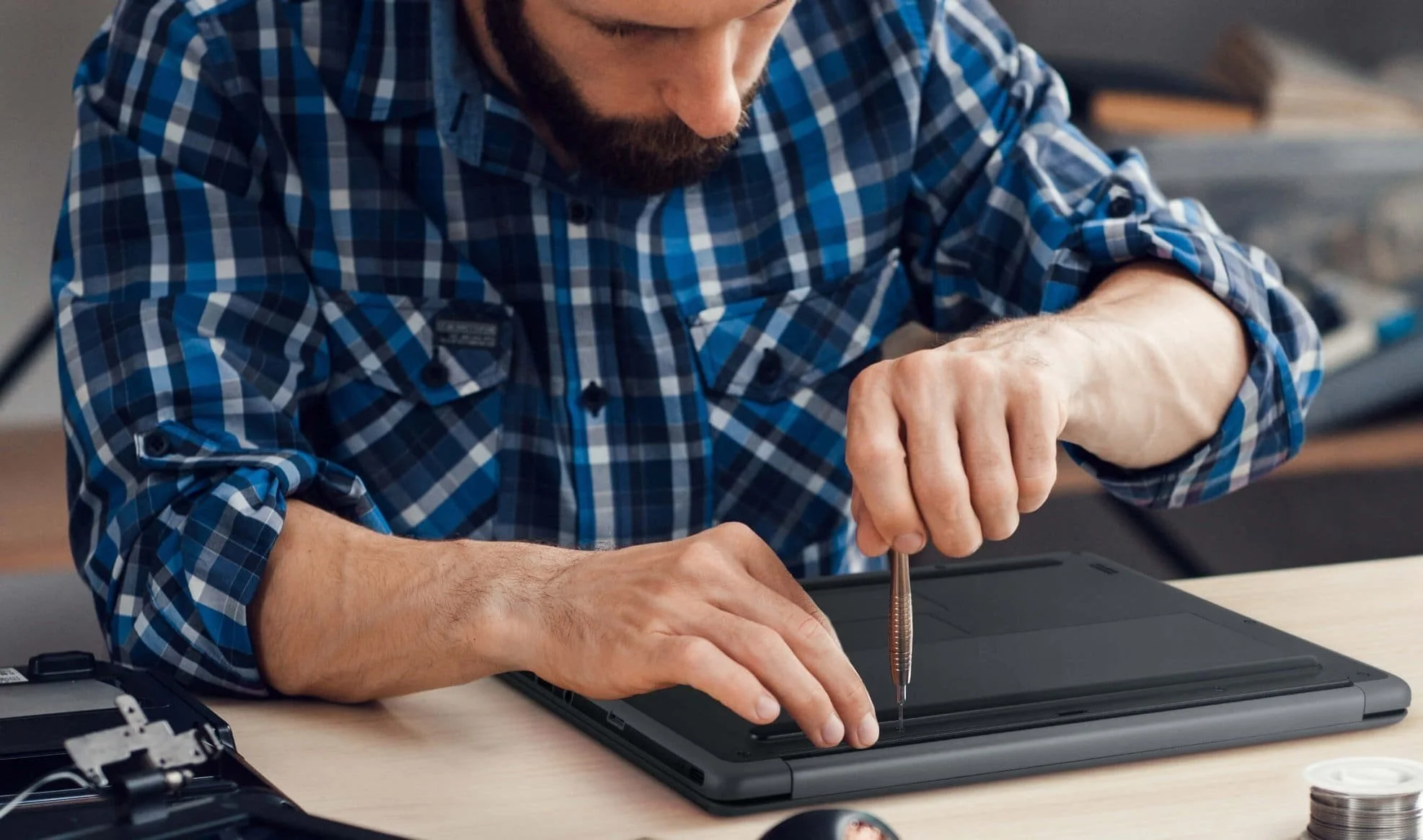 Person in blue plaid shirt repairing a laptop with a screwdriver on a wooden table.