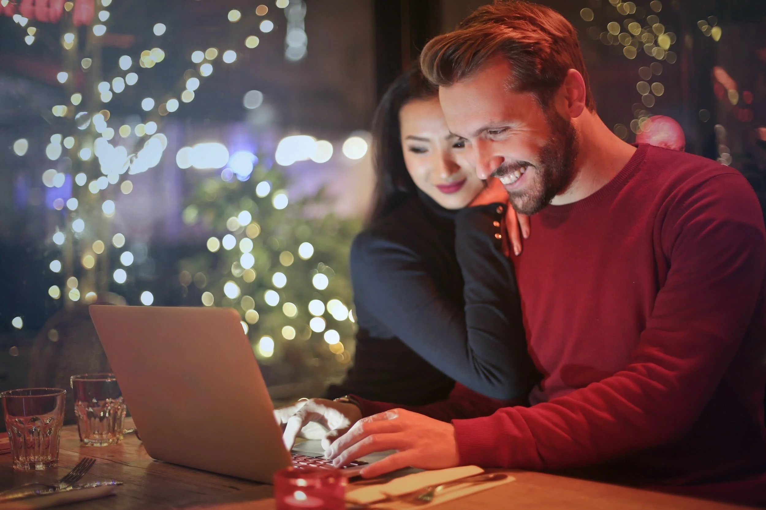 A smiling man and woman looking at a laptop screen together in a restaurant with dim lighting and fairy lights in the background.