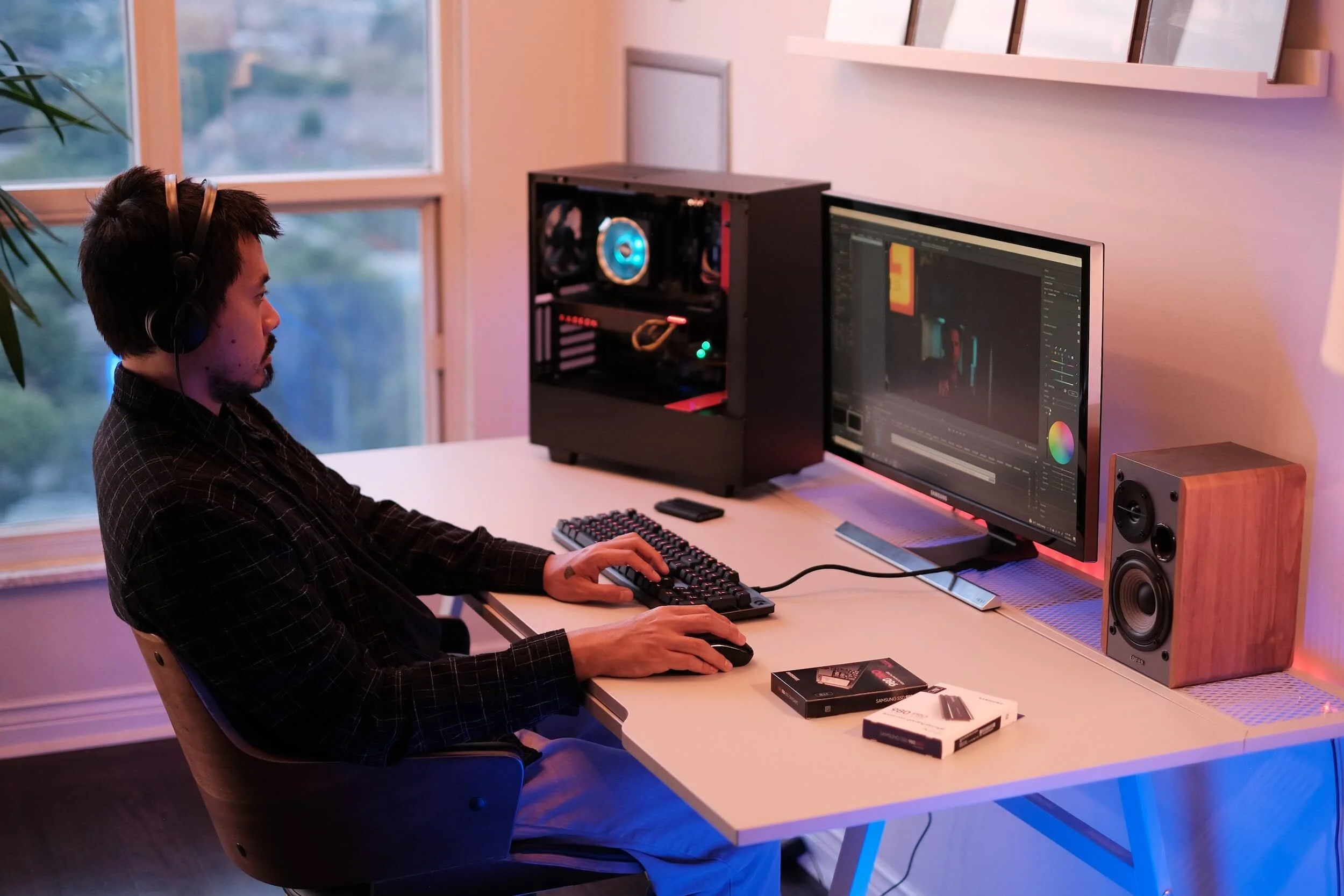 Person using a desktop computer, wearing headphones, seated at a desk with a monitor displaying video editing software. The desk also has a speaker and product boxes.