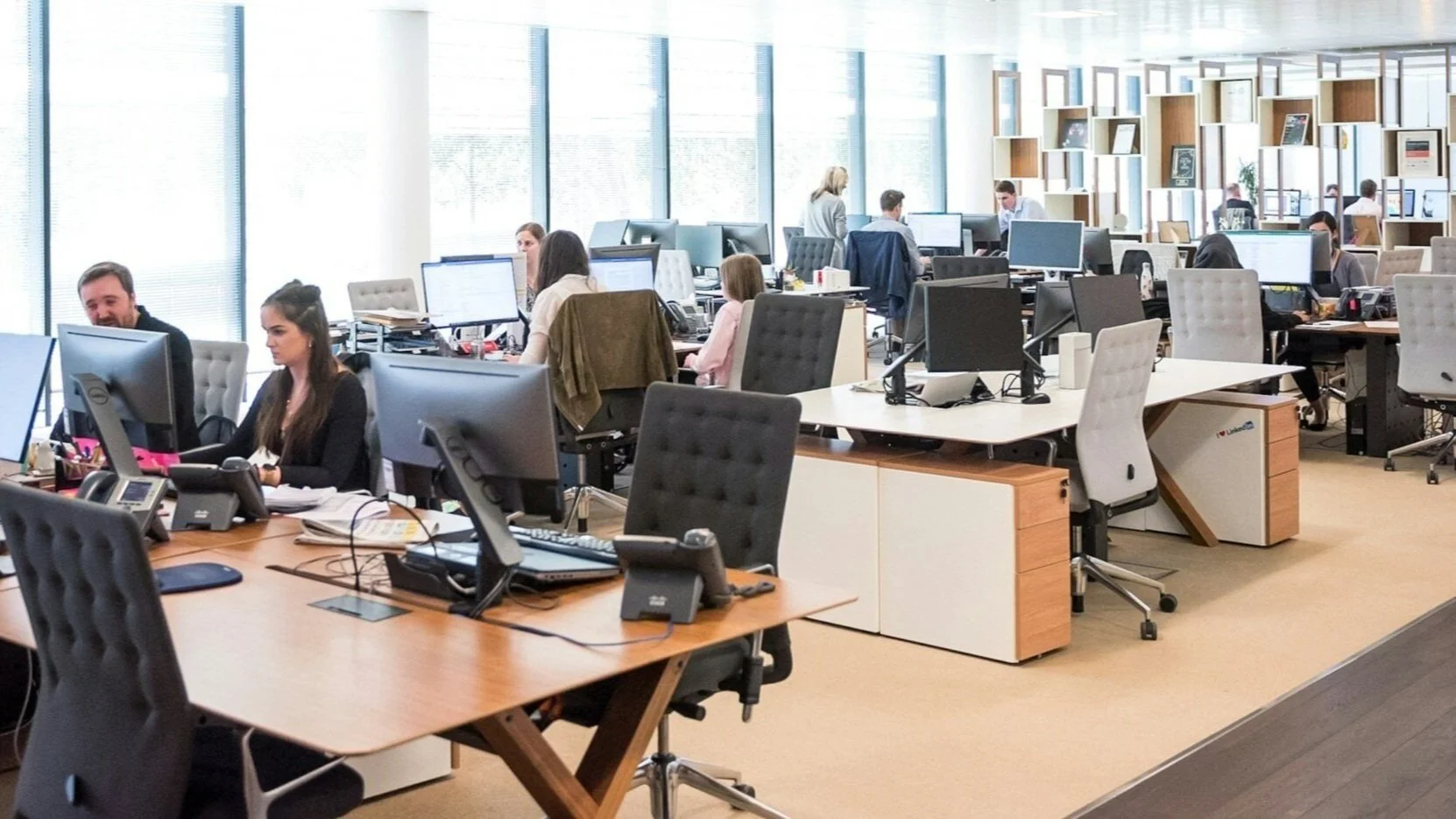 Office space with multiple employees working at desks with computers, large windows on one side, and a bookshelf in the background.