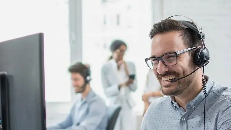 A man wearing glasses and a headset with a microphone, smiling while working at his computer. In the background, two colleagues are visible, also wearing headsets, in an office setting.
