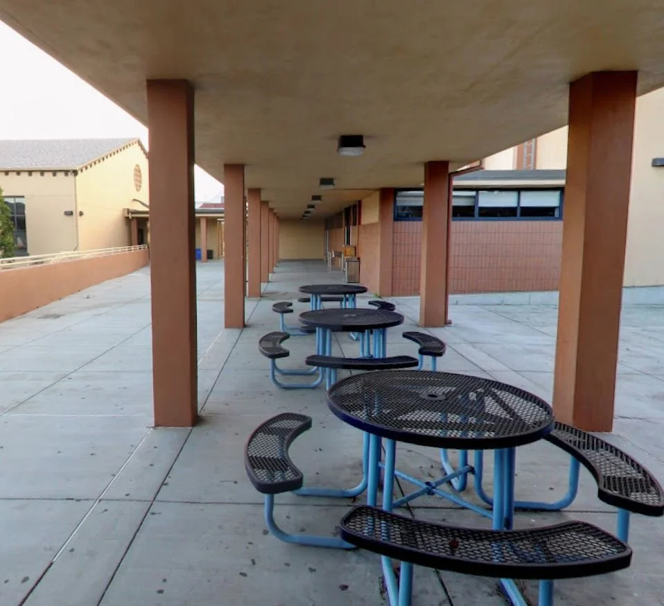 Empty outdoor area with a row of round picnic tables with attached benches under a covered walkway in front of a school building.