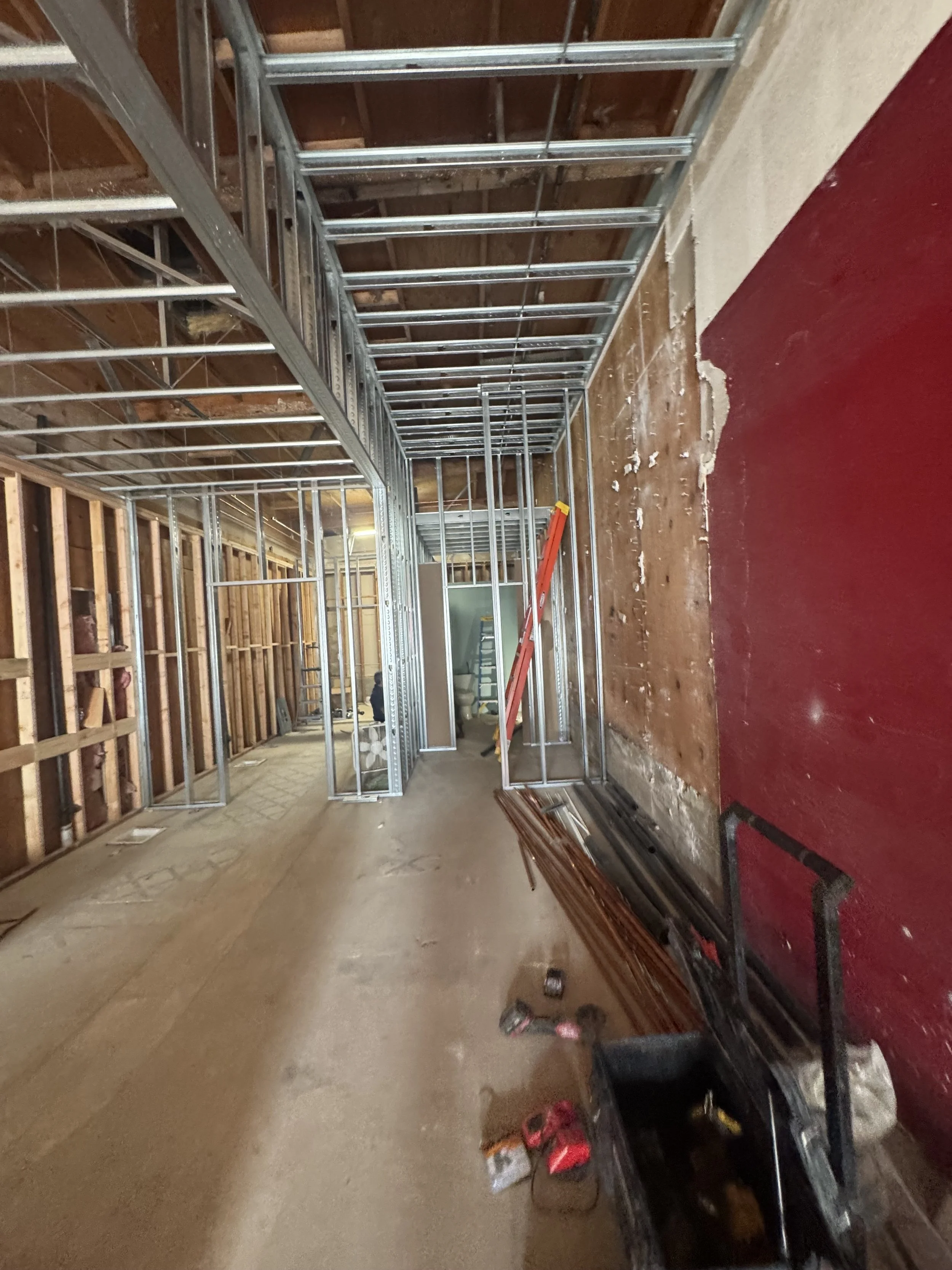 Interior view of a building under construction with exposed wall framing, metal studs, and ceiling framework. Construction tools and materials are scattered on the floor, and a ladder leans against the wall.