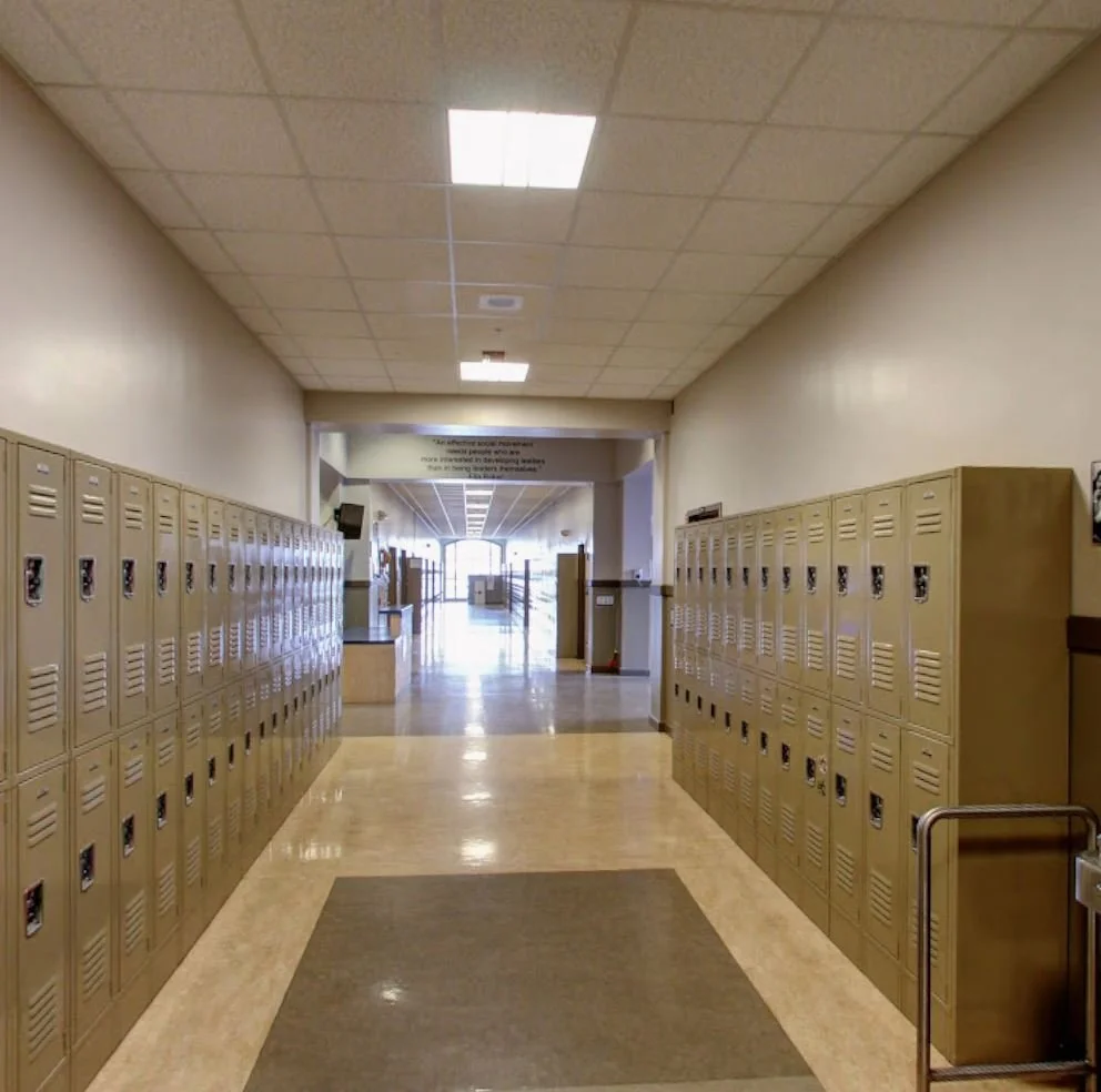 School hallway with beige lockers lining both sides, a tiled ceiling with fluorescent lights, and an exit at the end of the corridor with sunlight streaming through.