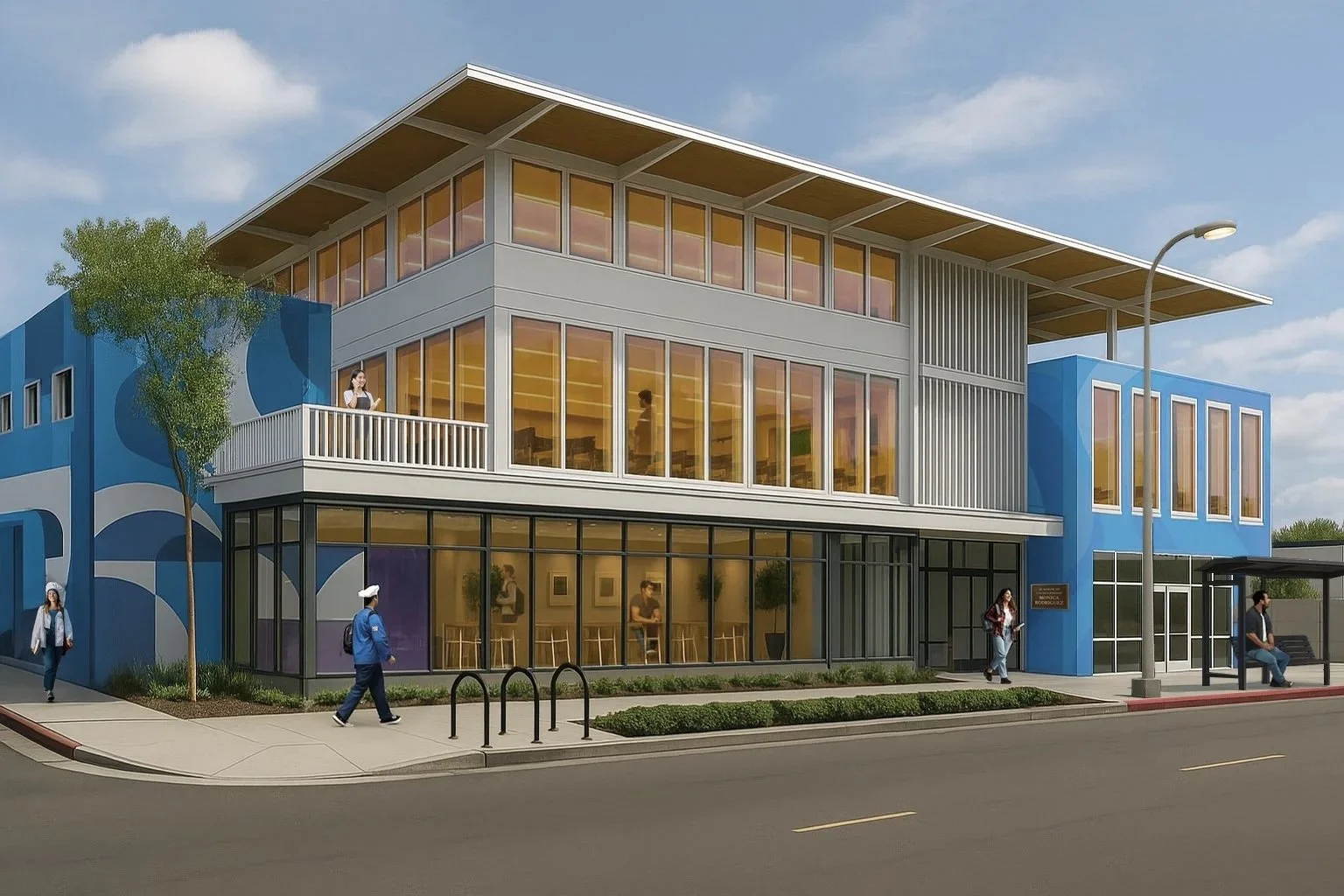 A modern, multi-story building with large glass windows, a blue and white exterior, and an outdoor balcony. People are walking on the sidewalk in front of it, and there is a bus stop with a bench and a streetlamp nearby.