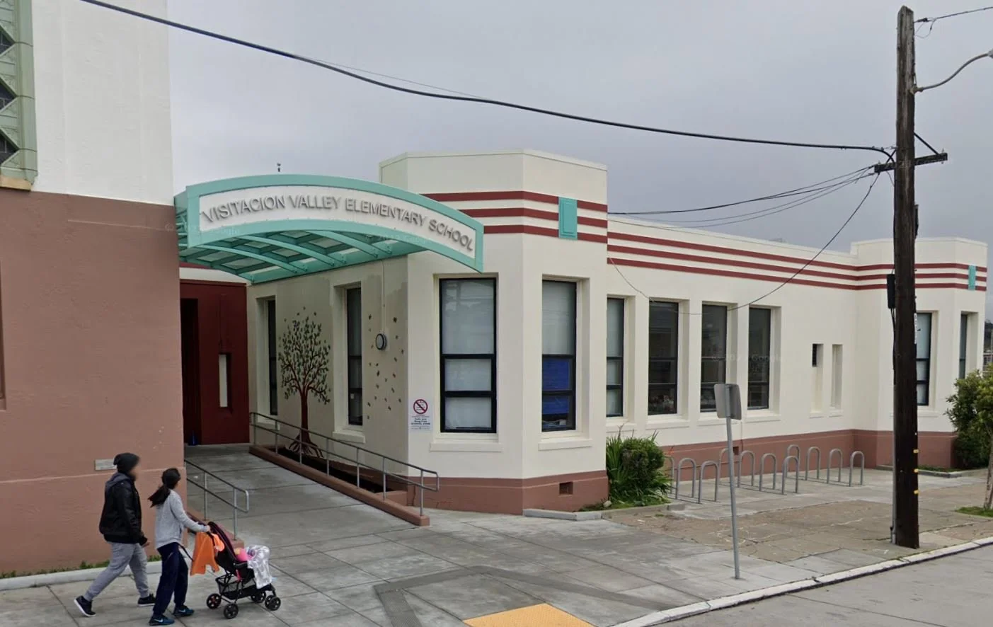 Exterior of Vistaon Valley Elementary School with three children walking on sidewalk, one pushing a stroller, and a building with a tree mural near the entrance.