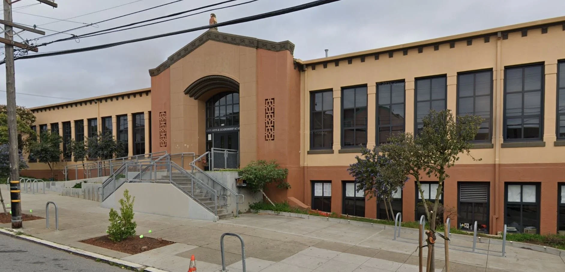 Exterior of a two-story school building with beige walls, large windows, and a main entrance with stairs and a ramp. The school is called City Arts & Leadership Academy.