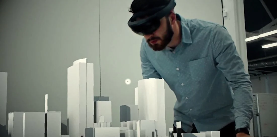 A man wearing a visor and gray shirt is examining a scale model of buildings on a table in an indoor space.