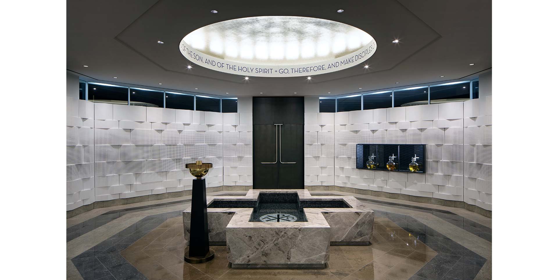 Interior view of a modern religious space with a central marble water fountain, decorative white textured walls, a black door, and a circular skylight with a religious inscription around it.