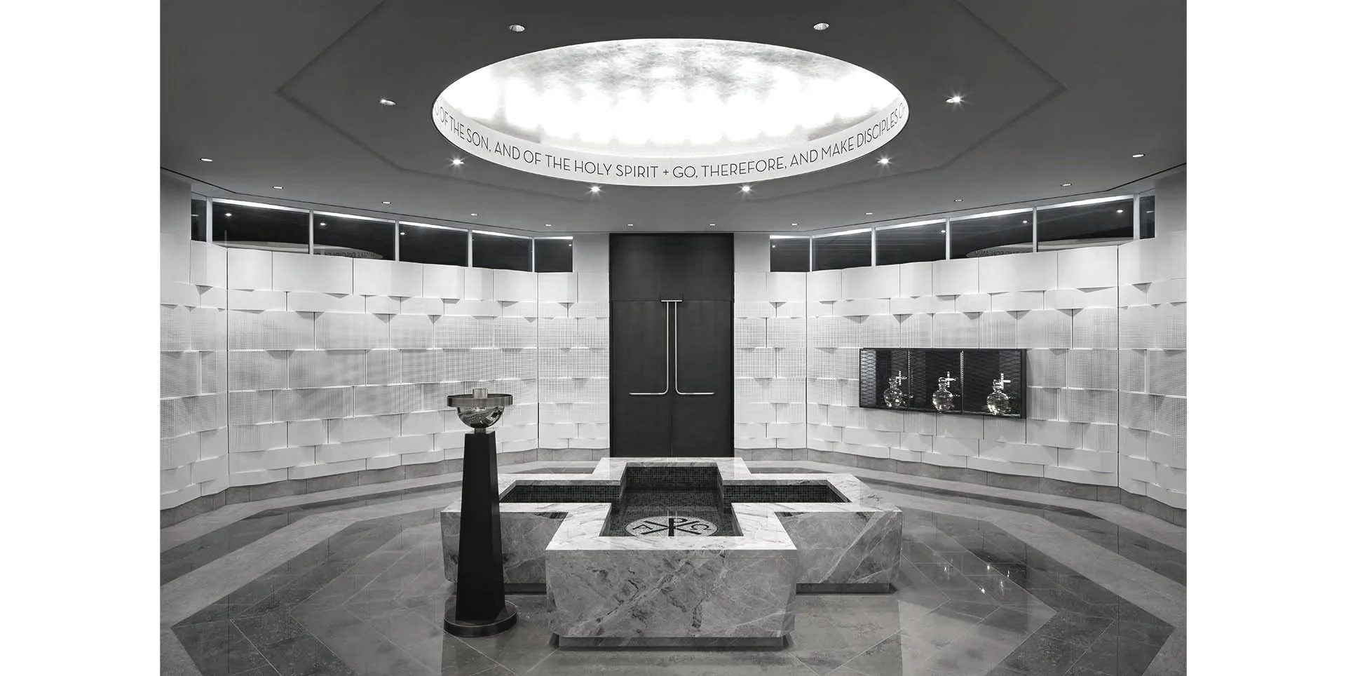 Interior view of a modern church with a circular marble baptismal font, a black door with a cross handle, and a sunken pool with a clock design, all surrounded by white textured walls and ceiling with a circular illuminated text reading 'OF THE SON, AND OF THE HOLY SPIRIT + GO, THEREFORE, AND MAKE DISCIPLES.'