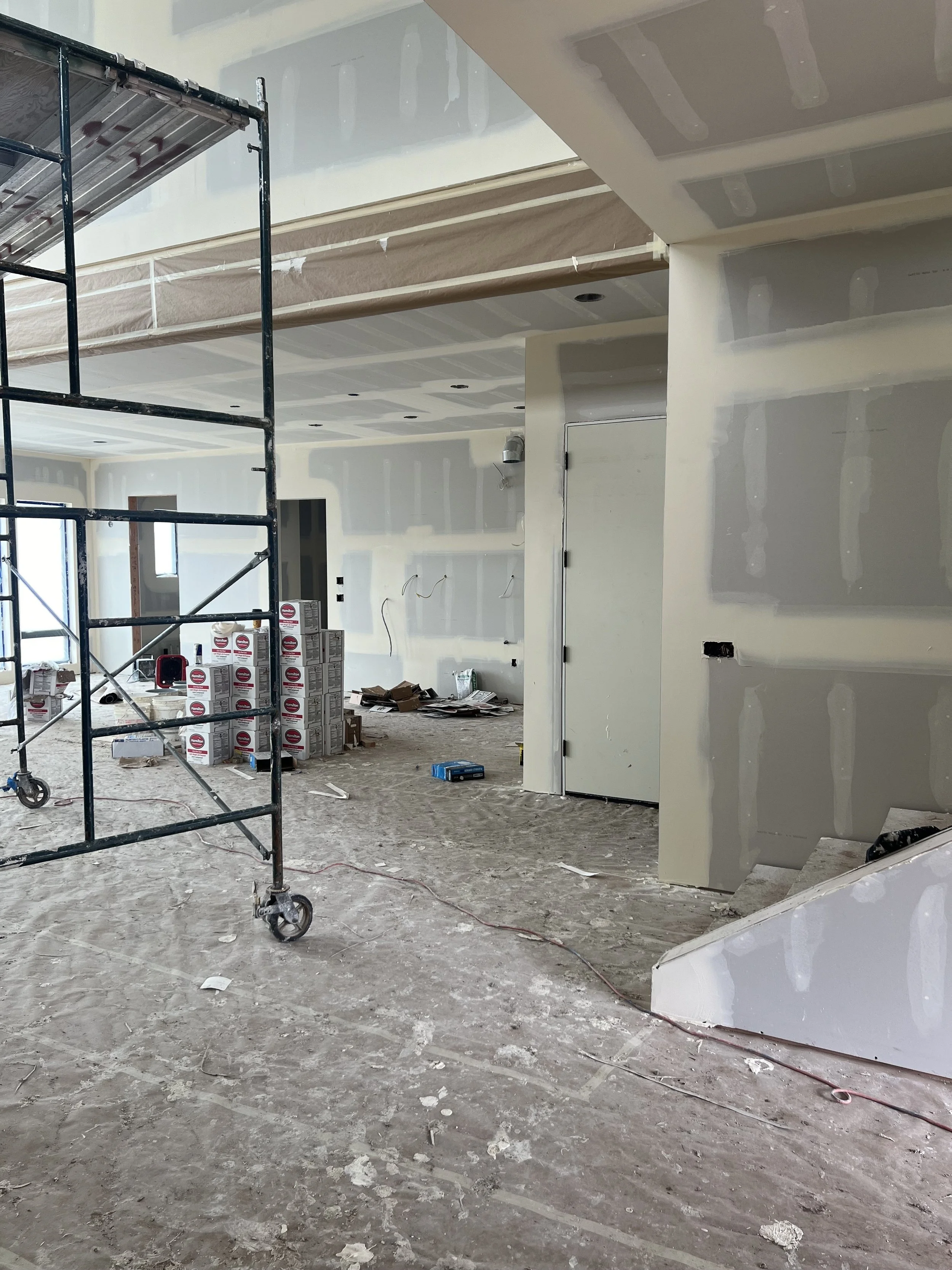 Interior of a building under construction, with drywall installation in progress, scaffolding on the left, and construction materials and tools scattered on the floor.