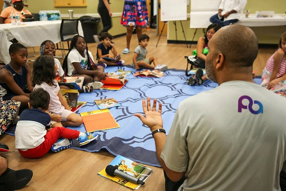 Diverse group of children seated in a circle with a man leading a reading activity.