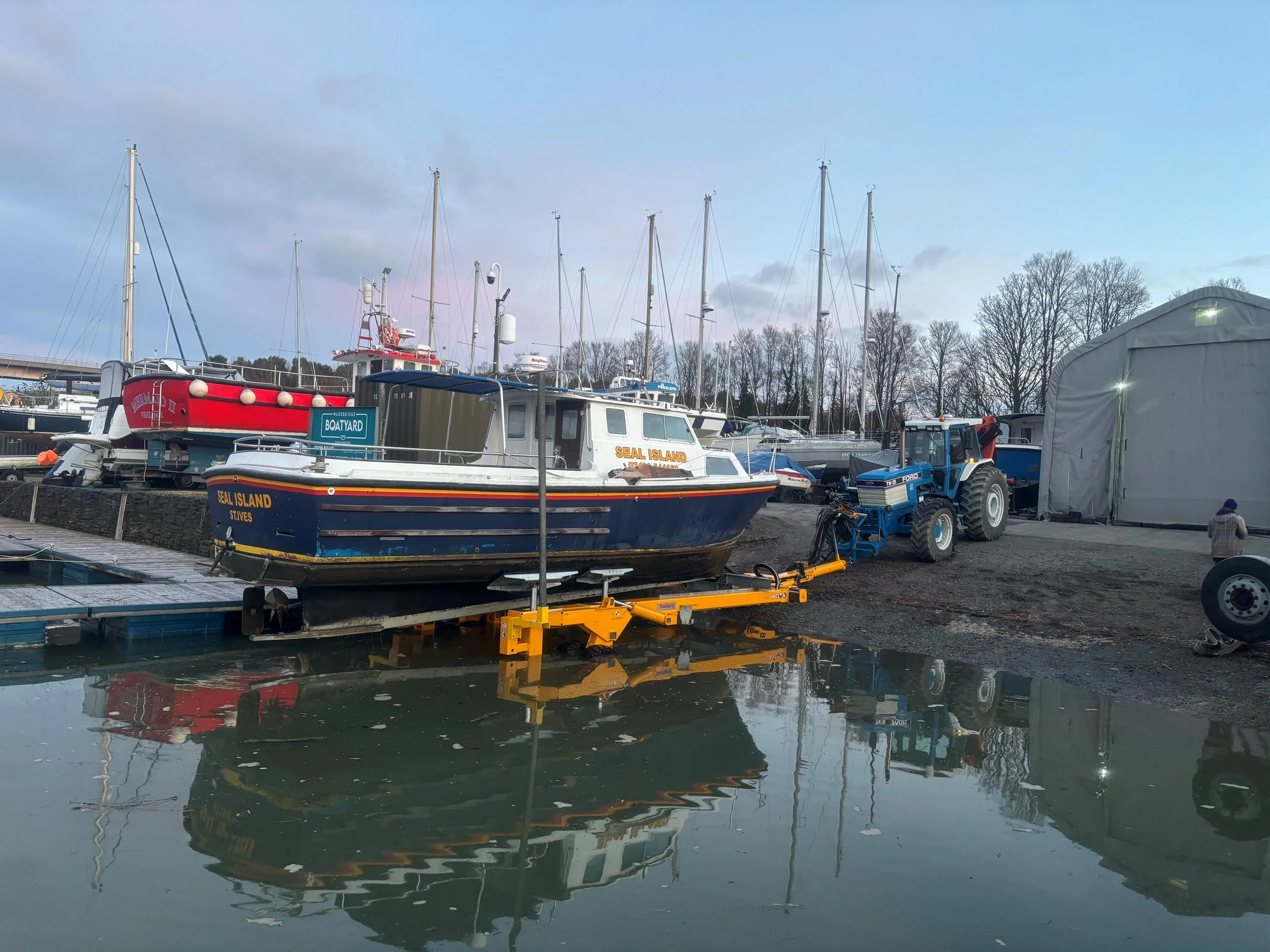 A boat with the name 'Seal Island' on the side is on a floating dock in a marina, surrounded by sailboats and a blue tractor, with trees and a cloudy sky in the background.