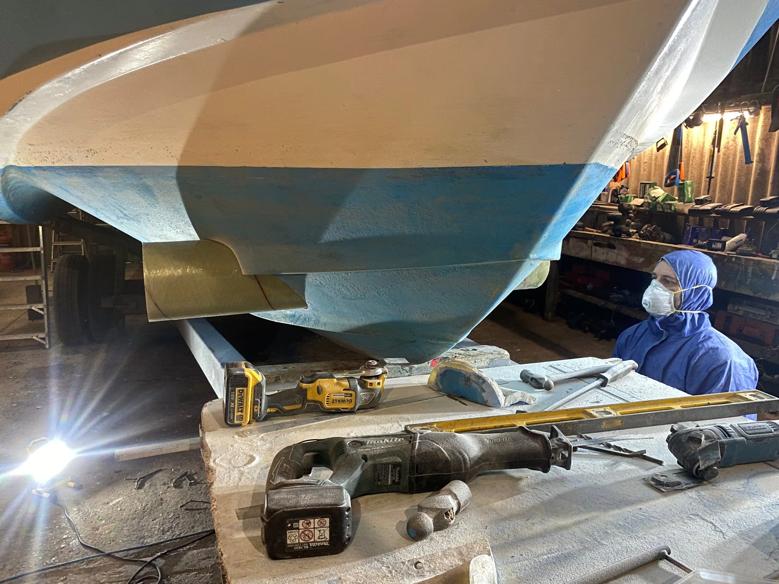 A person in blue protective gear and a face mask inspecting a boat in a workshop, with tools like drills and level on a workbench.
