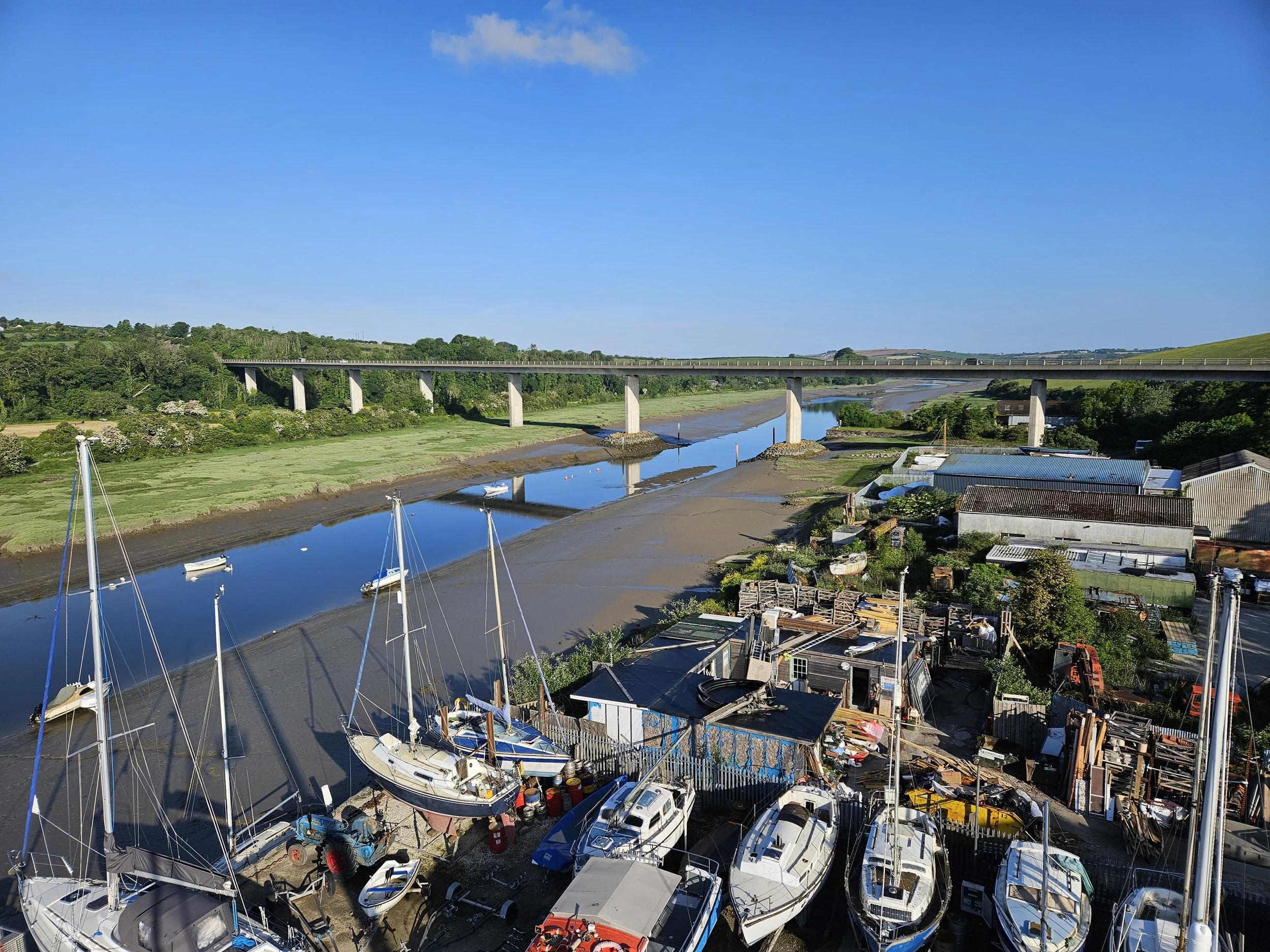 Sailboats docked in a marina with a river and green hills, a bridge spans the river in the background, under a clear blue sky.