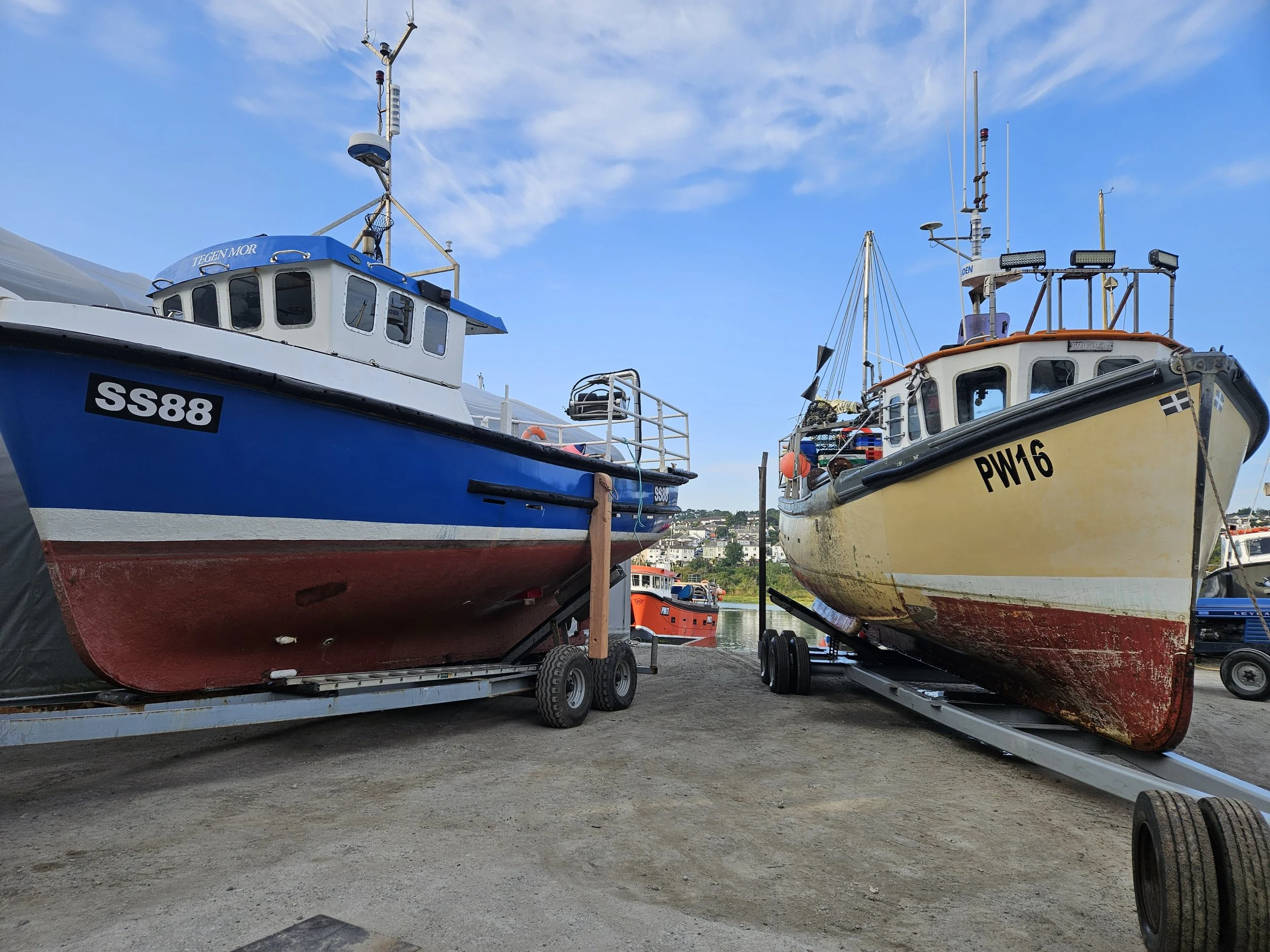 Two boats on trailers at a marina, one blue with the label 'SS88' and the other yellow with the label 'PW16', with water and buildings in the background under a partly cloudy sky.
