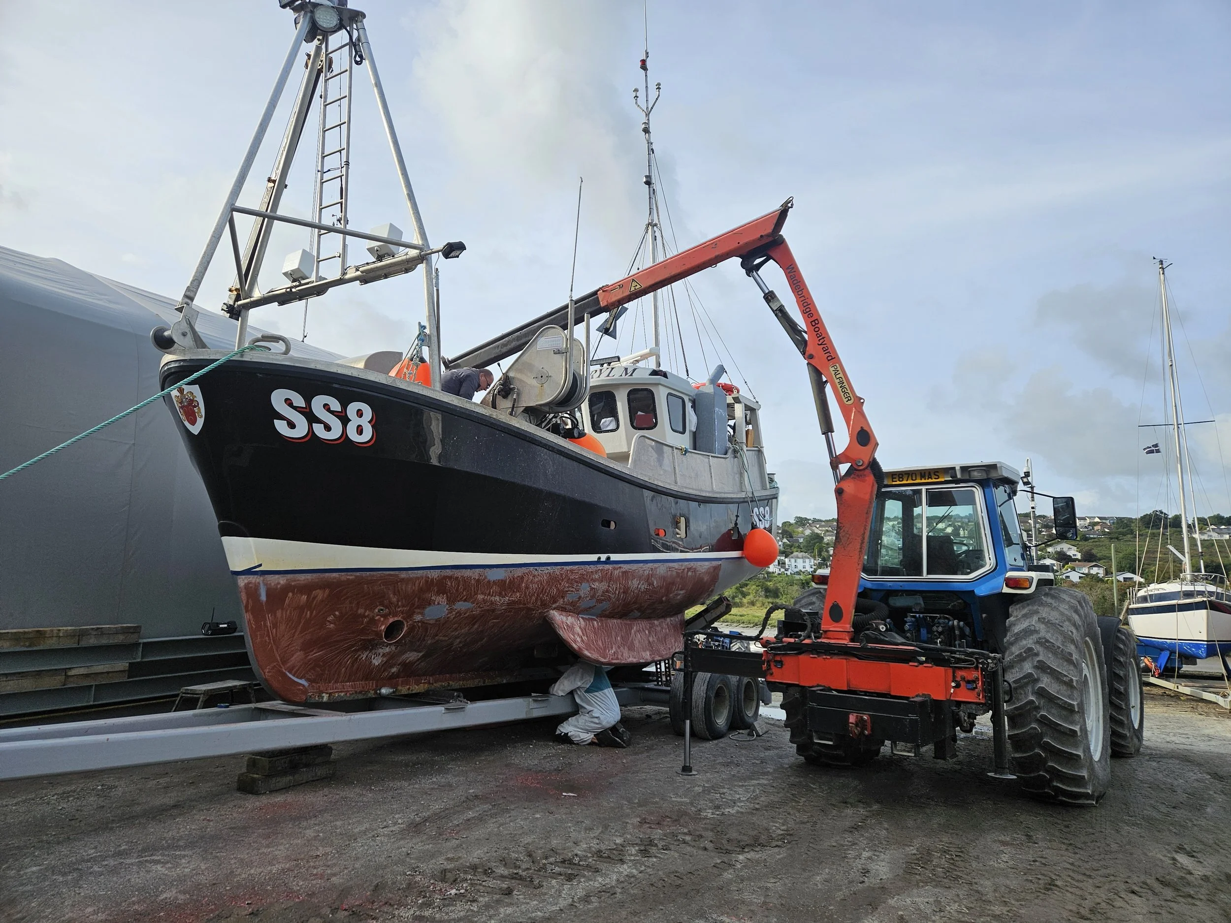 A boat on a trailer being lifted or moved by a blue and black tractor with a large arm and orange crane attachment at a boatyard or marina.